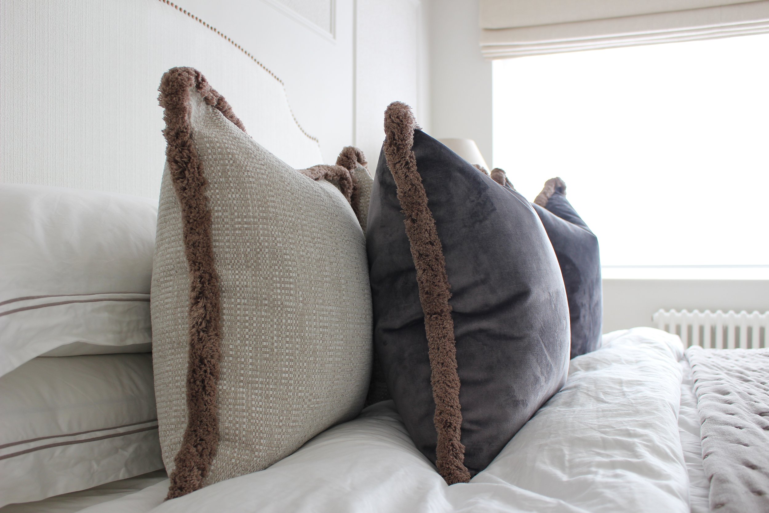 Close-up of decorative throw pillows with brown fringe edges on a bed with white sheets and pillows, sunlight coming through a window. Master bedroom designed by Yazzmin Lovelle Interiors