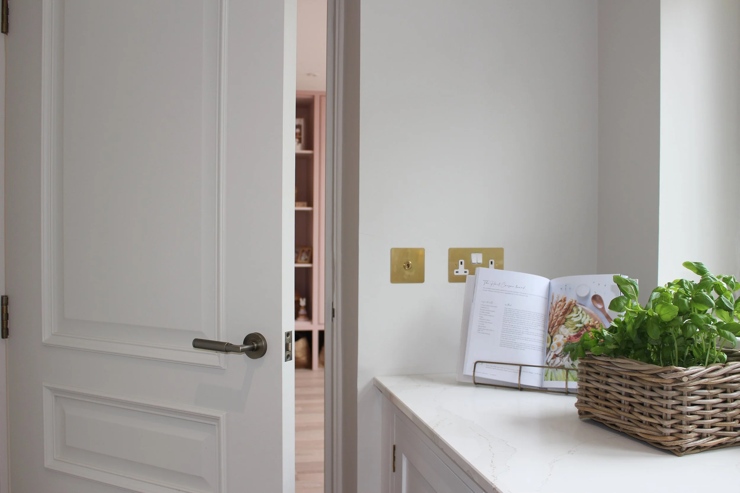 A white interior door is slightly open, revealing another room with pink walls and a shelf. Beside the door, a white countertop or table holds an open magazine or cookbook in a black holder and a basket with a green plant.