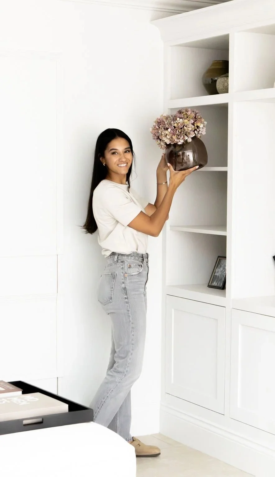 Woman placing a pink hydrangea bouquet in a dark brown vase on a media unit in a bright, traditionally styled room.