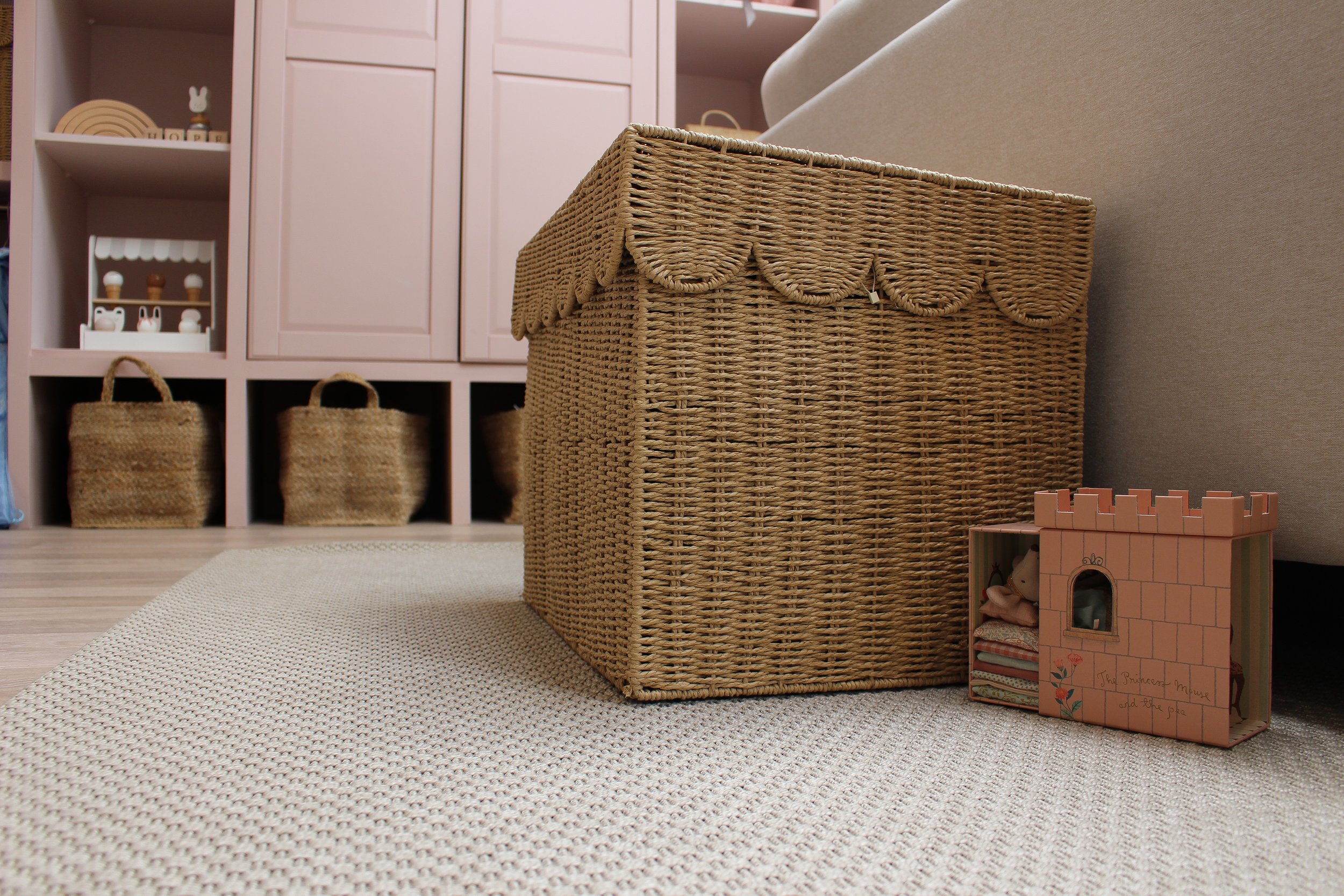 Children's playroom with a woven basket on a rug, storage unit with baskets and toys in the background, and a small toy castle on the floor. Playroom designed by Yazzmin Lovelle Interiors.