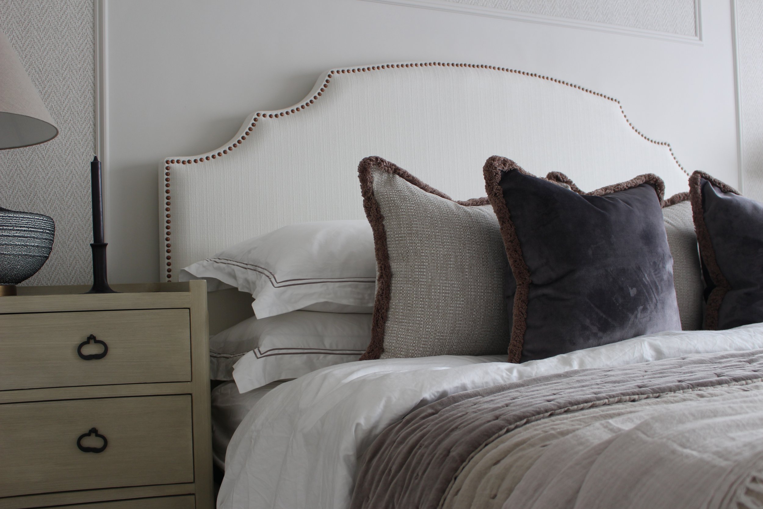 A neatly made bed featuring white linens and several decorative pillows in various textures and colours, with a cream-coloured headboard, next to a light wood nightstand with a lamp and decorative bowl. 