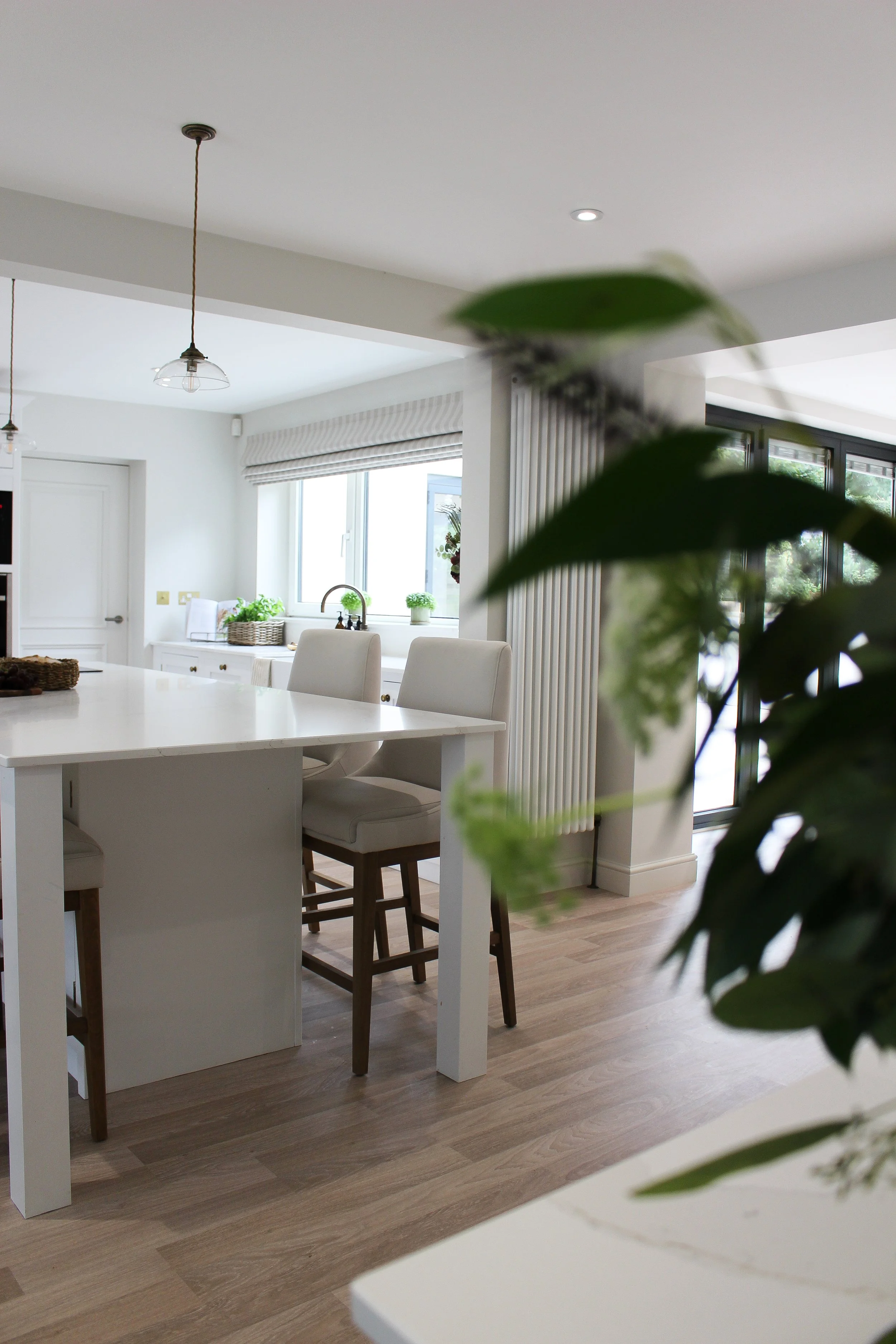 Modern kitchen with white island, light wood flooring, and large windows with blinds, partially obscured by a plant in the foreground.