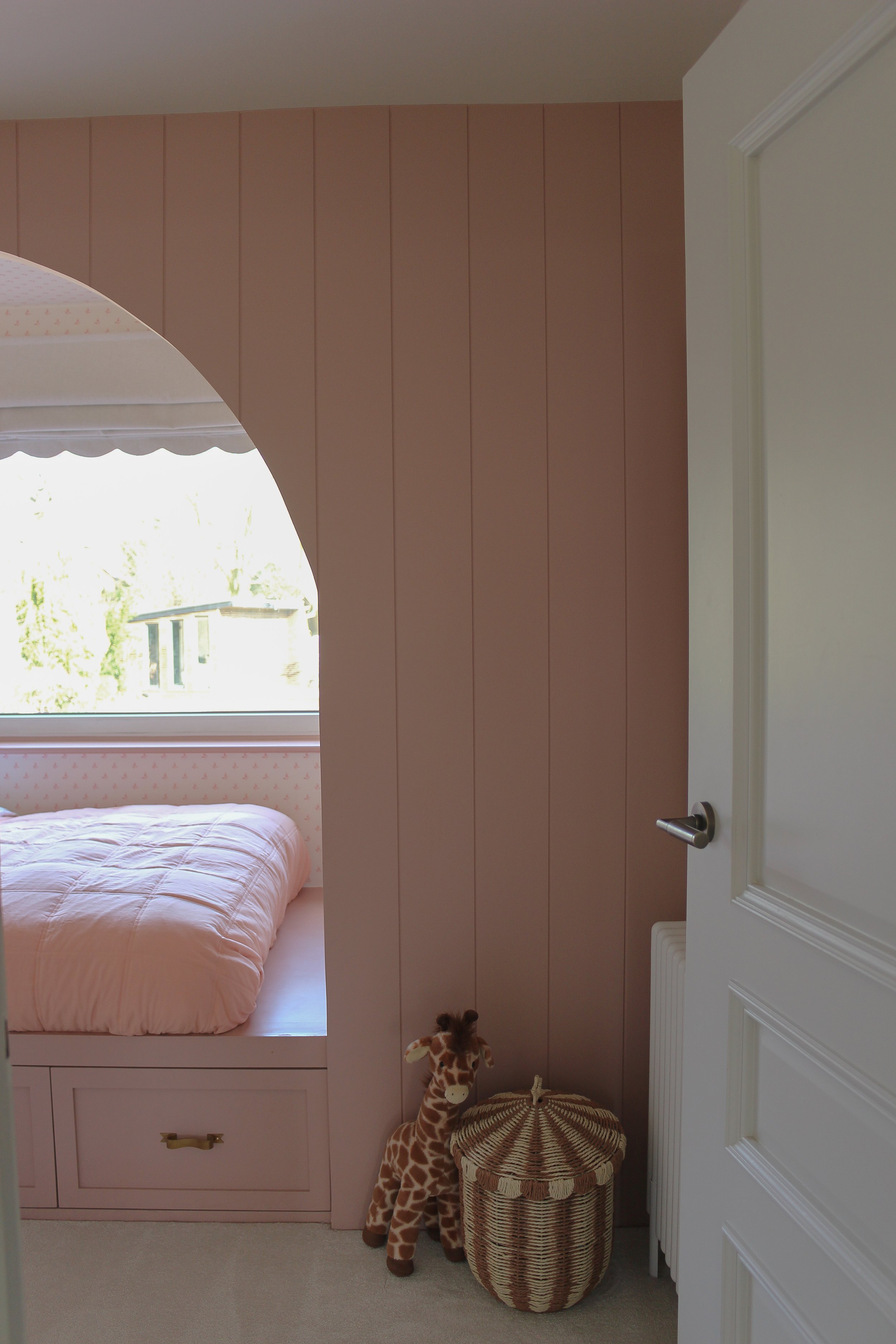A pink and beige children’s bedroom with a bed, toy giraffe, and woven storage basket near a door. Girls bedroom designed by Yazzmin Lovelle Interiors 