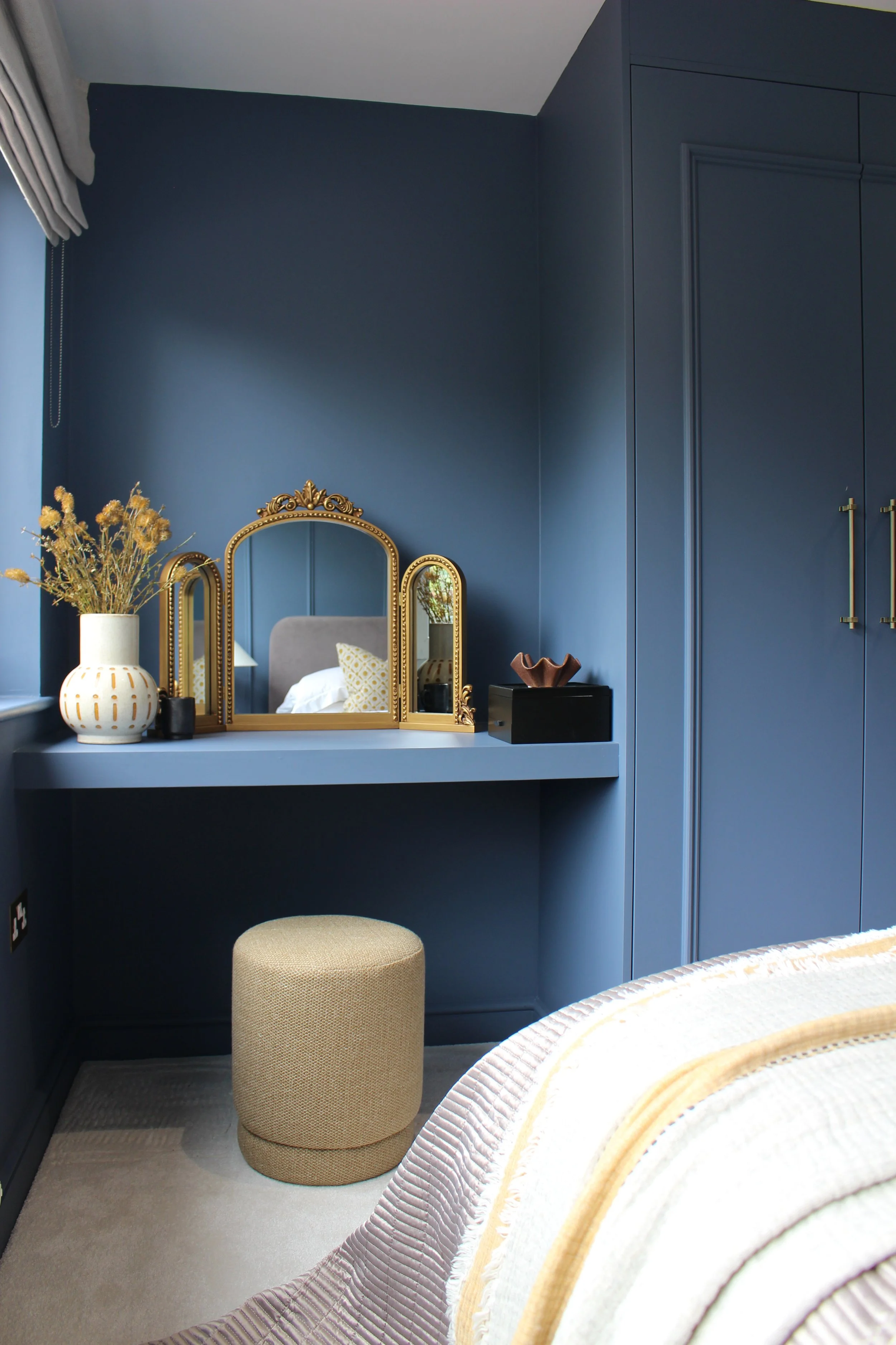 A bedroom corner with a blue walls, a vanity table with a gold framed mirror, a mustard ottoman, and decorative items including a vase with dried flowers. Guest bedroom designed by Yazzmin Lovelle Interiors.