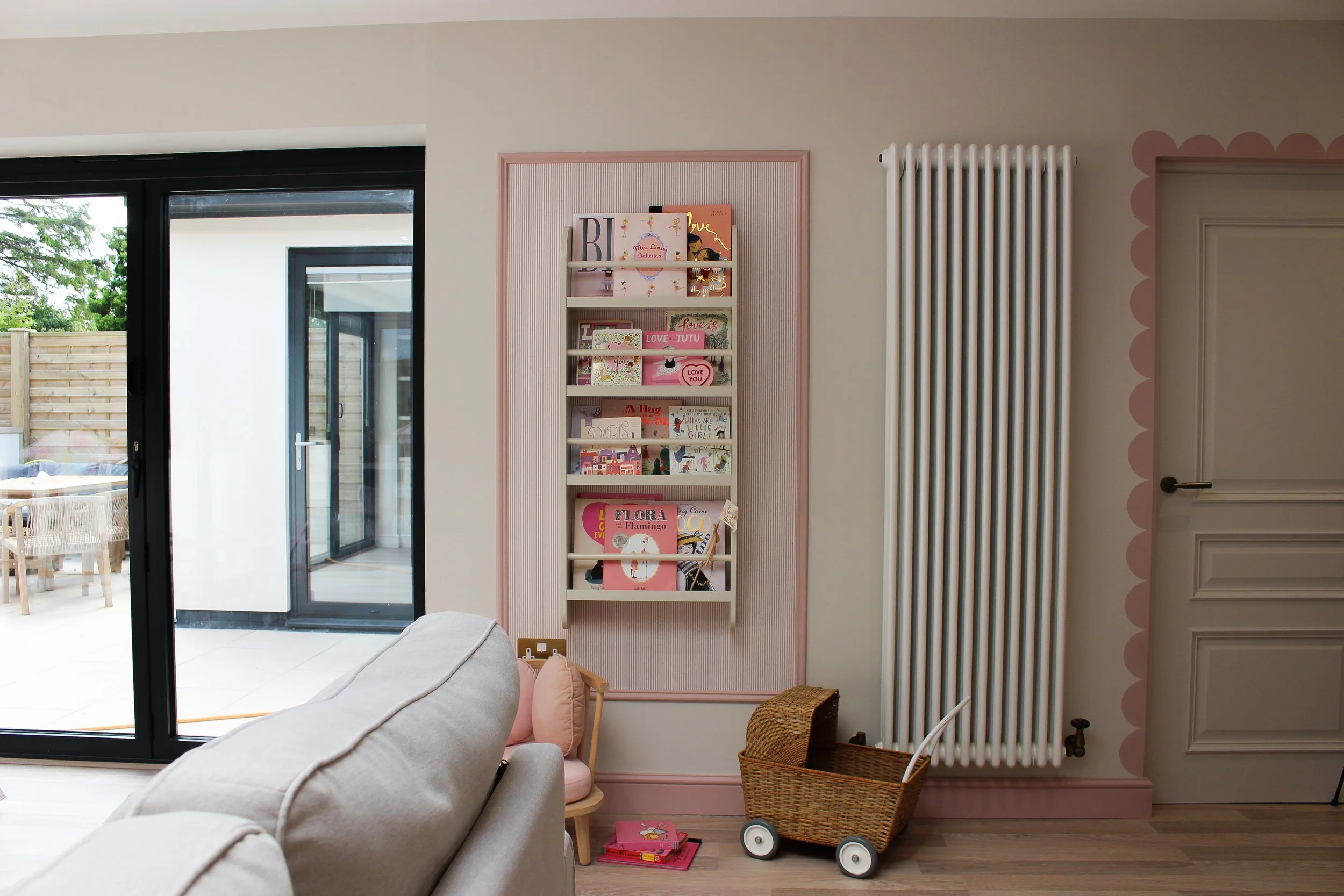 Playroom with pink and white bookshelf filled with books, wicker pram, beige sofa, and sliding glass door leading to a patio. painted scallop detail around door. Playroom designed by Yazzmin Lovelle Interiors.