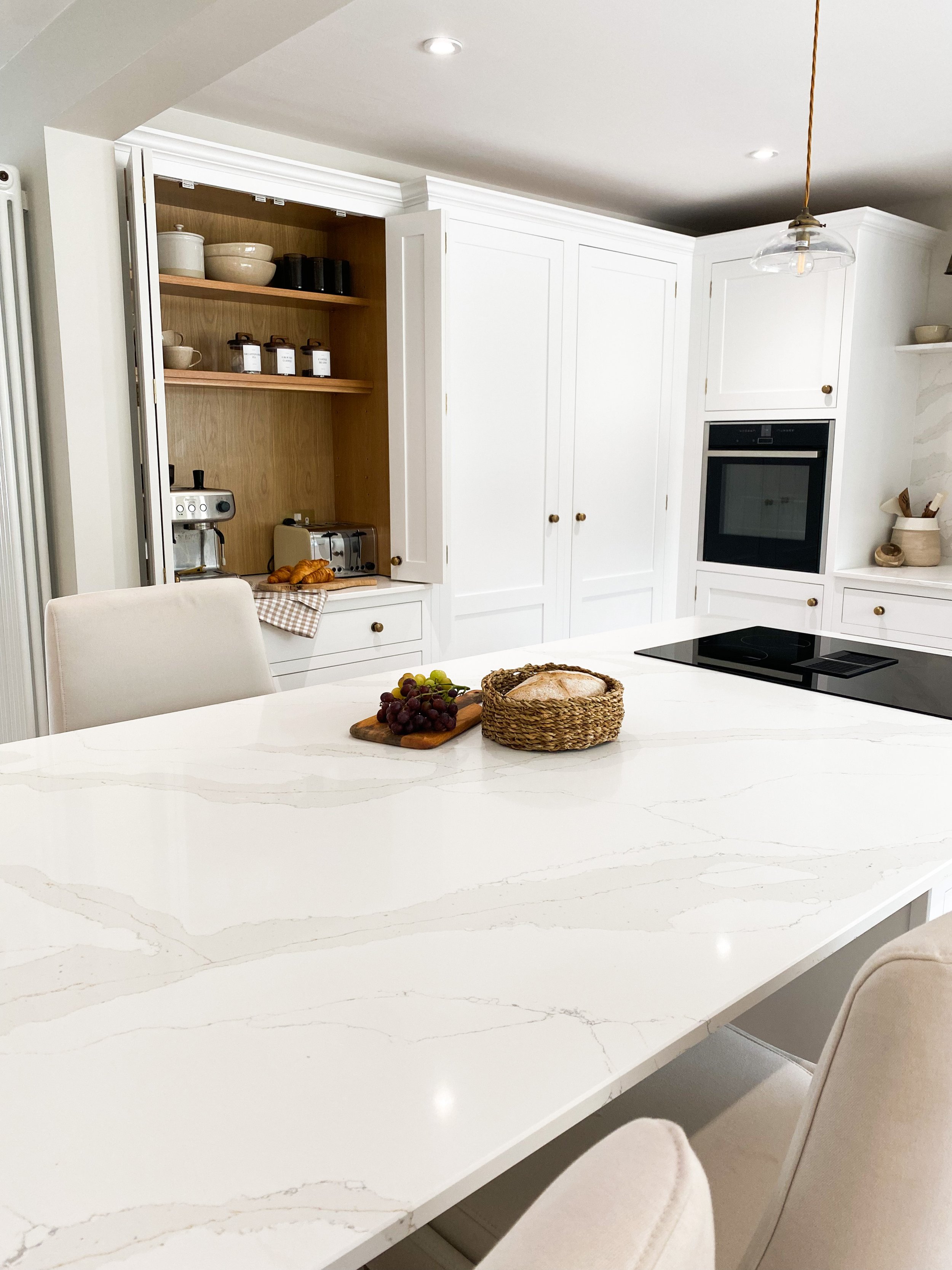 Modern kitchen with white cabinetry, a large white marble island, and minimalist decor. On the island, there are a bunch of grapes, a basket of bread, and a wooden cutting board. In the background, there are built-in shelves with kitchen items, a cof