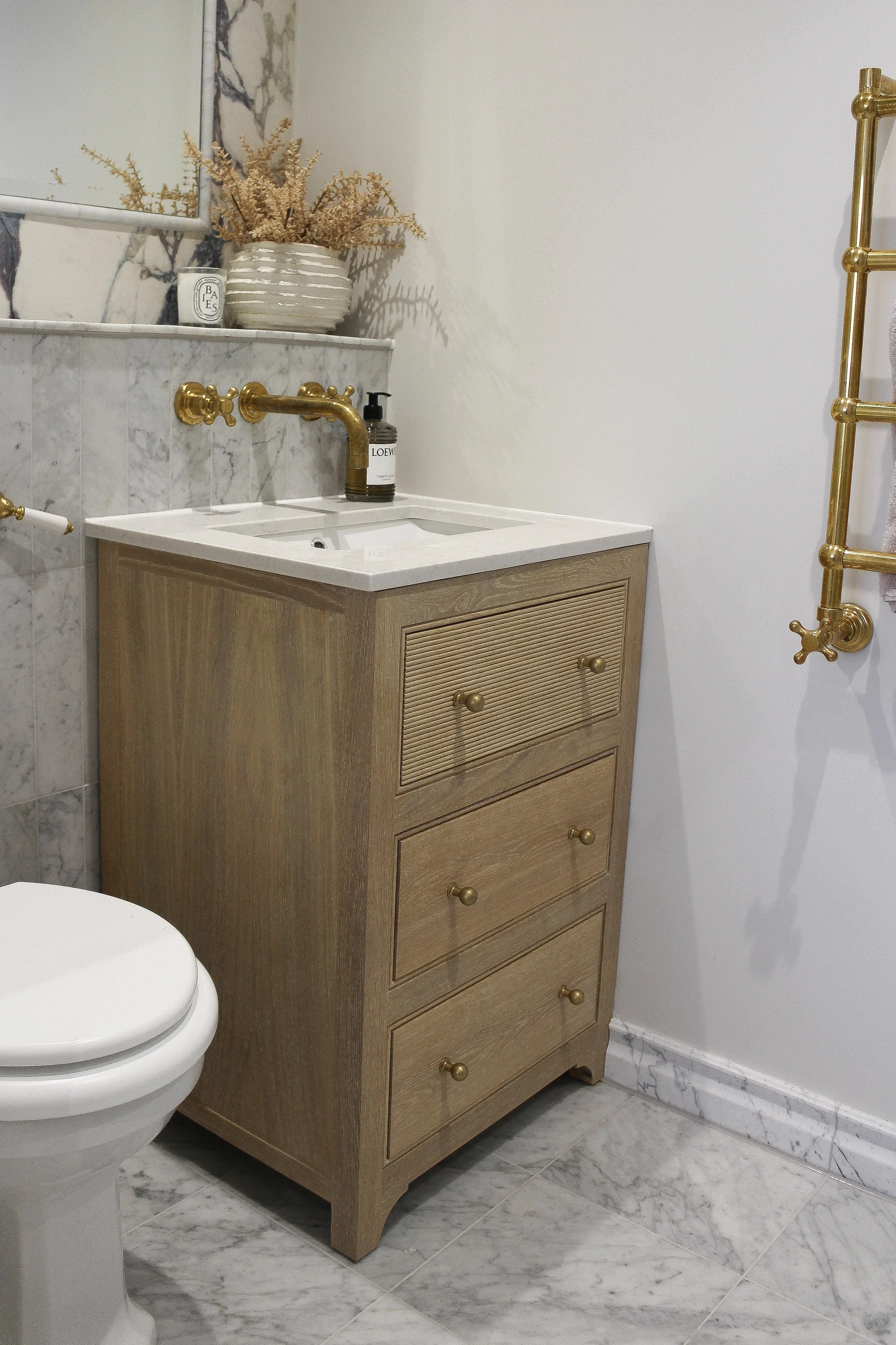 A Parker and Howley wooden bathroom vanity unit, next to a white traditional toilet in a ensuite bathroom with a marble tiled floor. Master ensuite designed by Yazzmin Lovelle Interiors