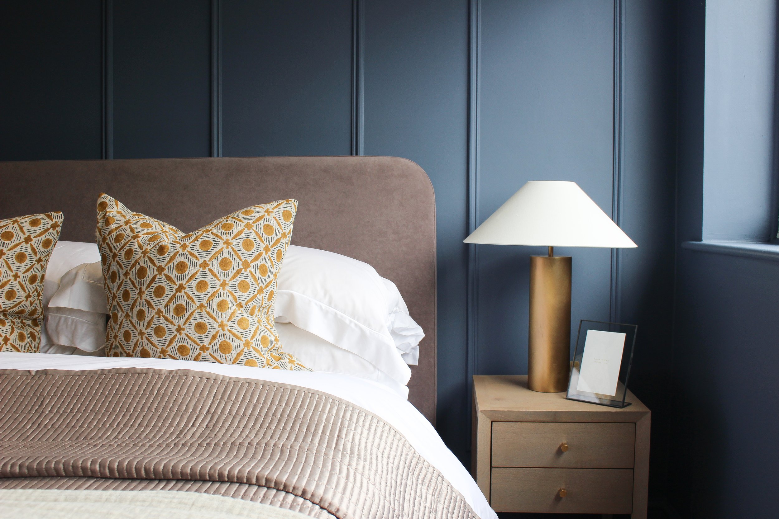 A neatly made bed with patterned throw cushions, white bedding and grey bed spread beside a wooden bedside table with a modern gold lamp and framed photo, against a dark blue panelled wall. Guest bedroom designed by Yazzmin Lovelle Interiors 