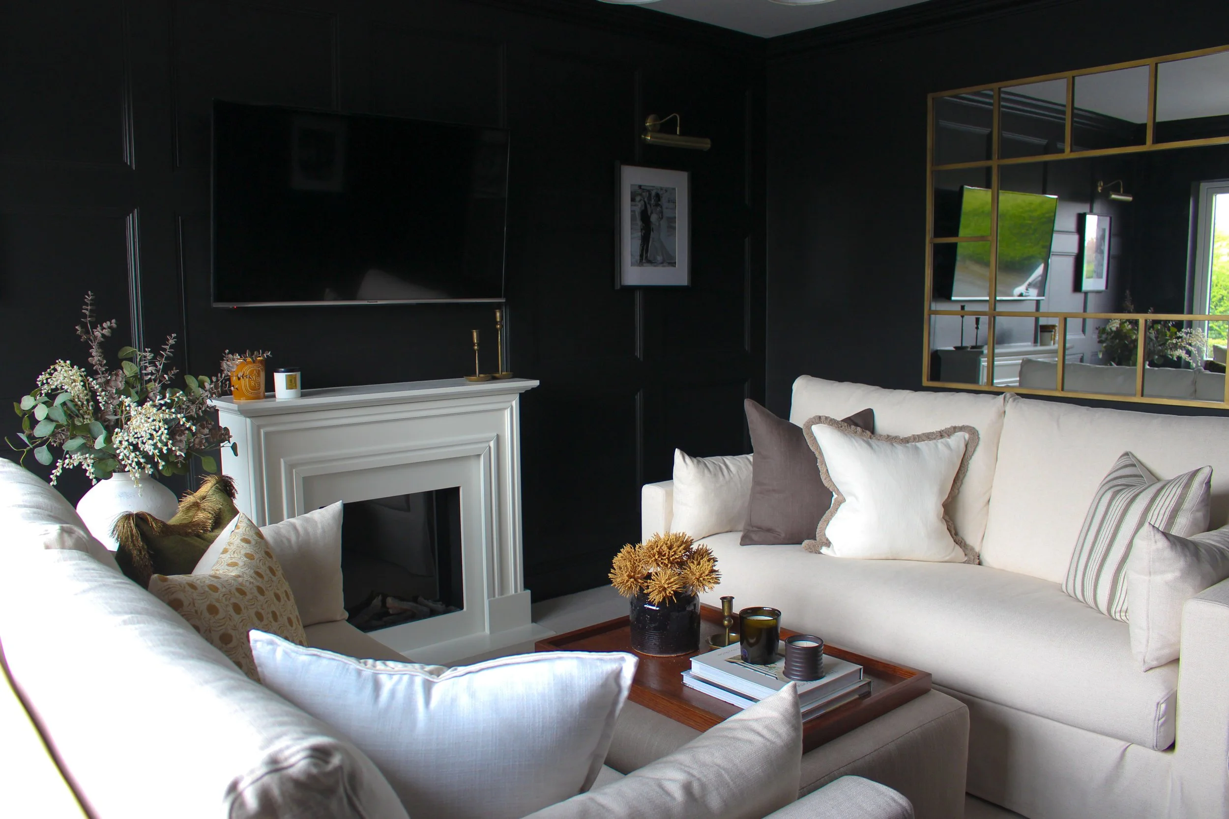 A living room with dark walls, a large white sofa with various pillows, a ottoman coffee table with decorative items, a white fireplace with decorative items on top, a mirror reflecting part of the room, and large windows allowing natural light.