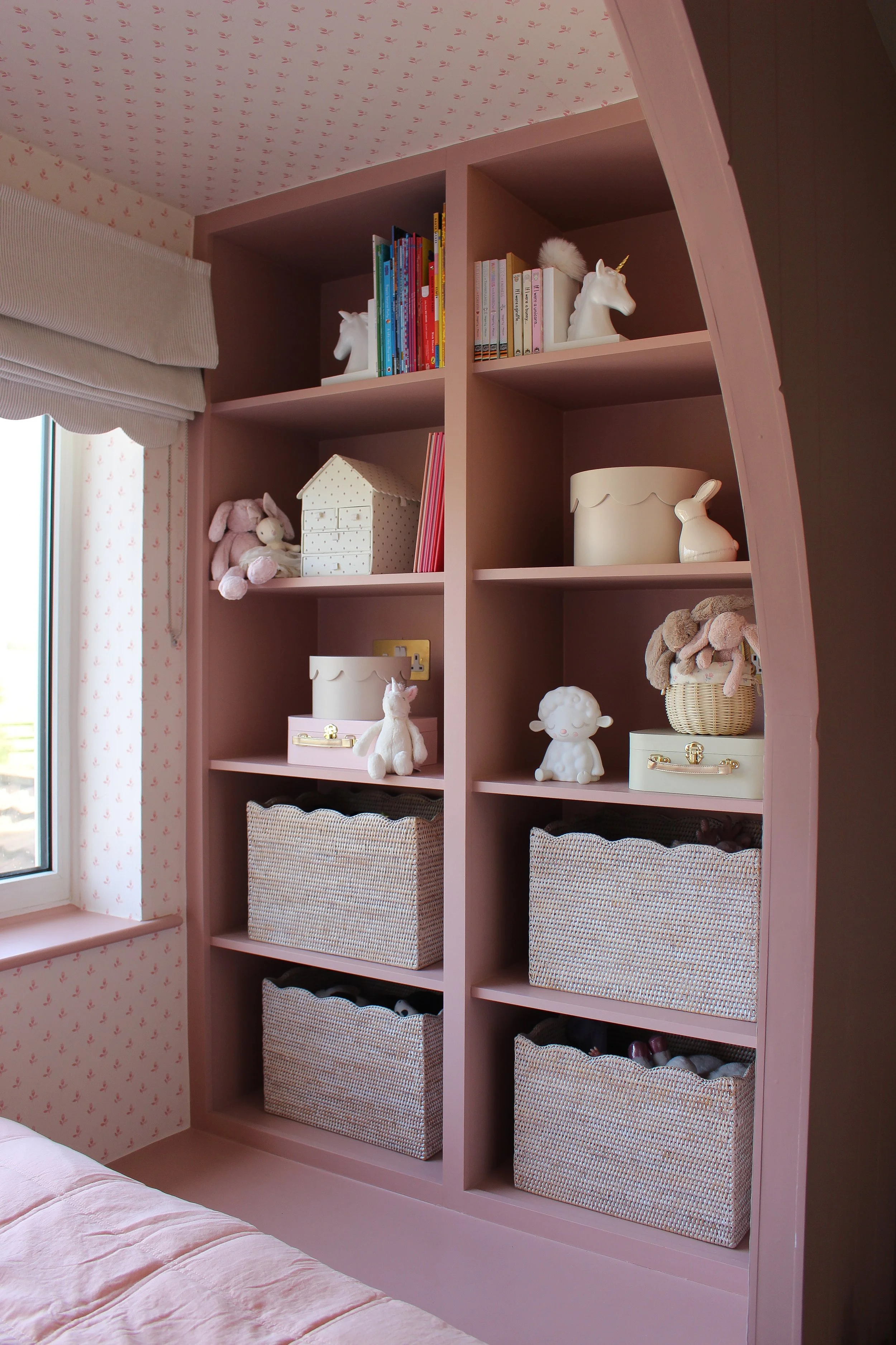 A pink wooden bookshelf in a children's bedroom filled with stuffed animals, decorative boxes, and books, next to a window with floral wallpaper. Girls bedroom designed by Yazzmin Lovelle Interiors