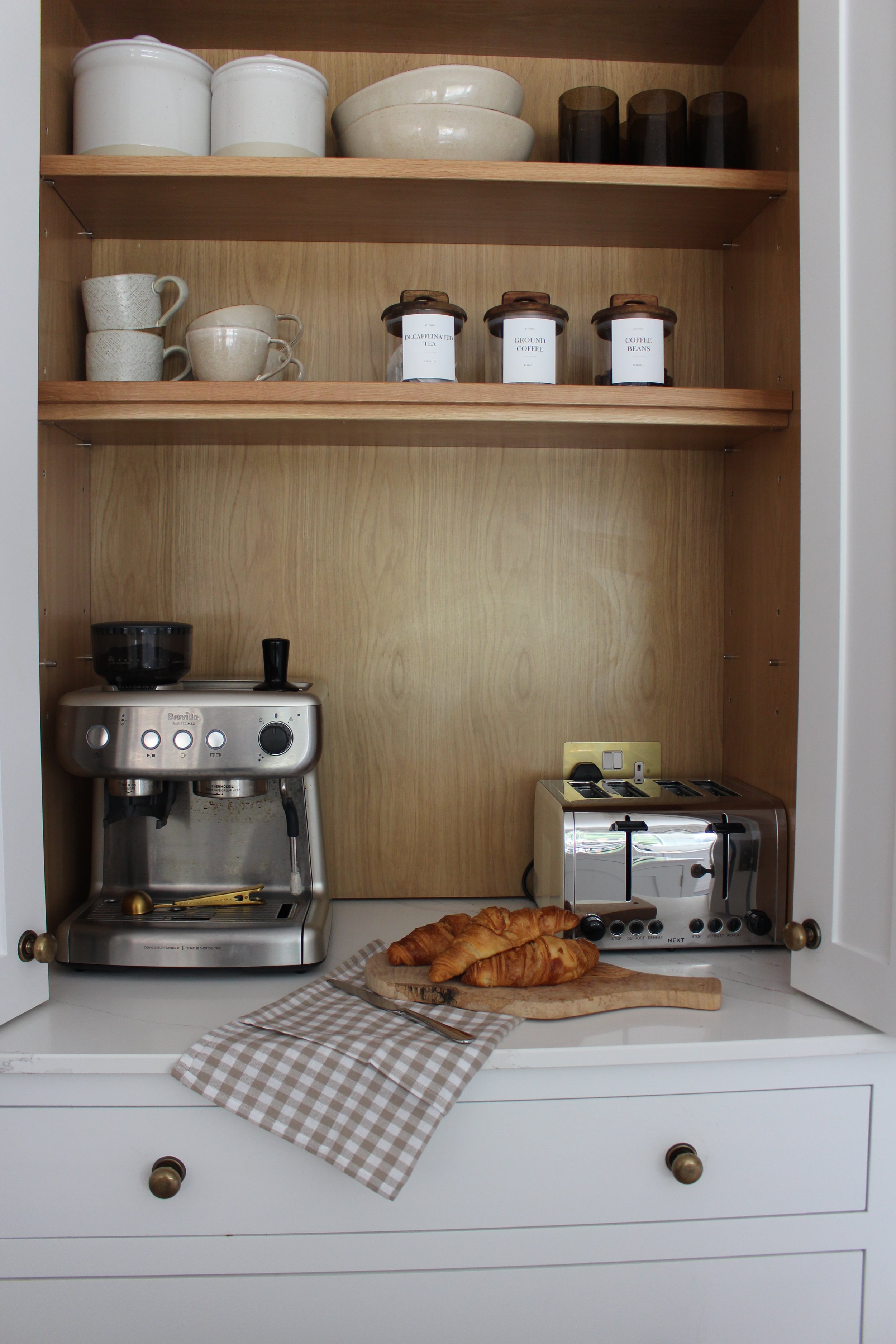 Open kitchen breakfast pantry cabinet with coffee machine, toaster, croissants on a cutting board, and mugs and jars on the shelves in a traditional kitchen from a yazzmin lovelle interiors project