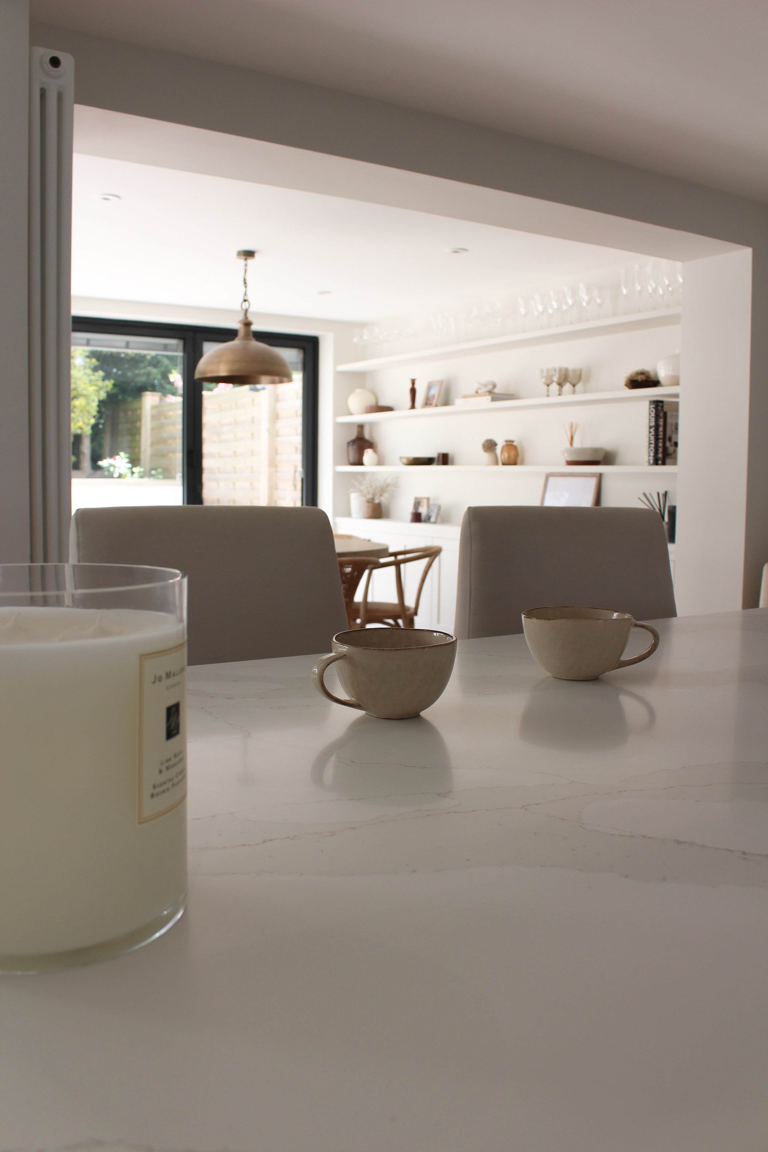 A dining table with two beige cups and a candle in a glass container, in a modern, well-lit kitchen with white walls and open shelving in the background.