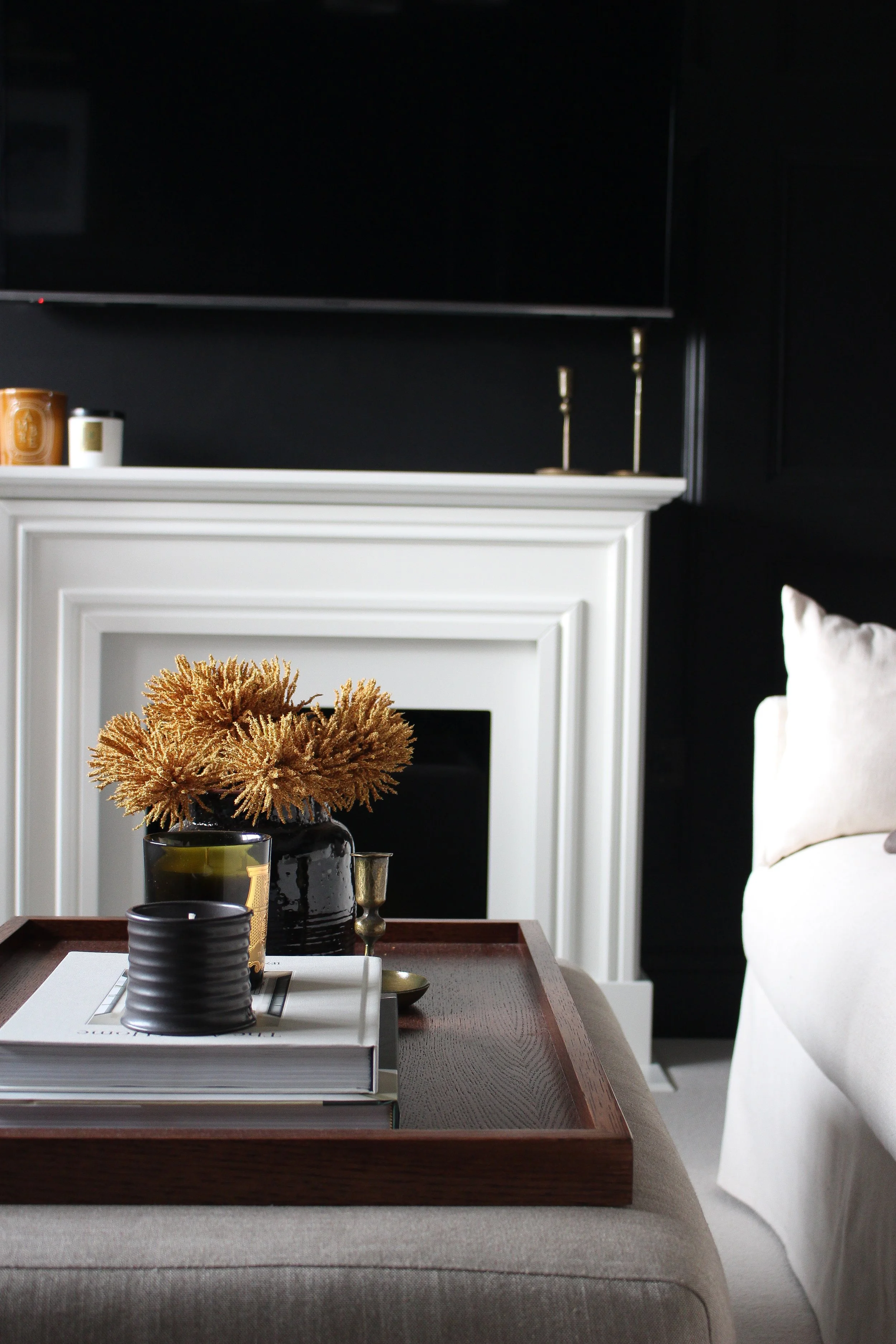 Living room with a white fireplace, beige sofa, wooden coffee table tray with books, black and gold decorative items, and a black vase with dried amber coloured plants. Black panelled walls. Living room designed by Yazzmin Lovelle Interiors.