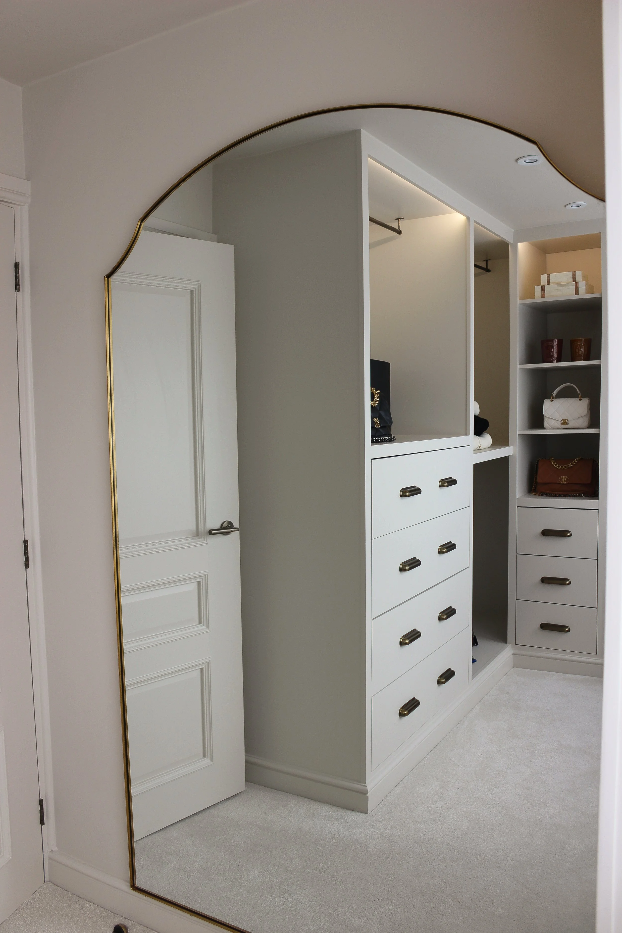 A large, gold framed mirror leaning against a wall in a walk in dressing room with off white shelves and drawers, containing handbags, boxes, and decorative items. Dressing room designed by Yazzmin Lovelle Interiors.
