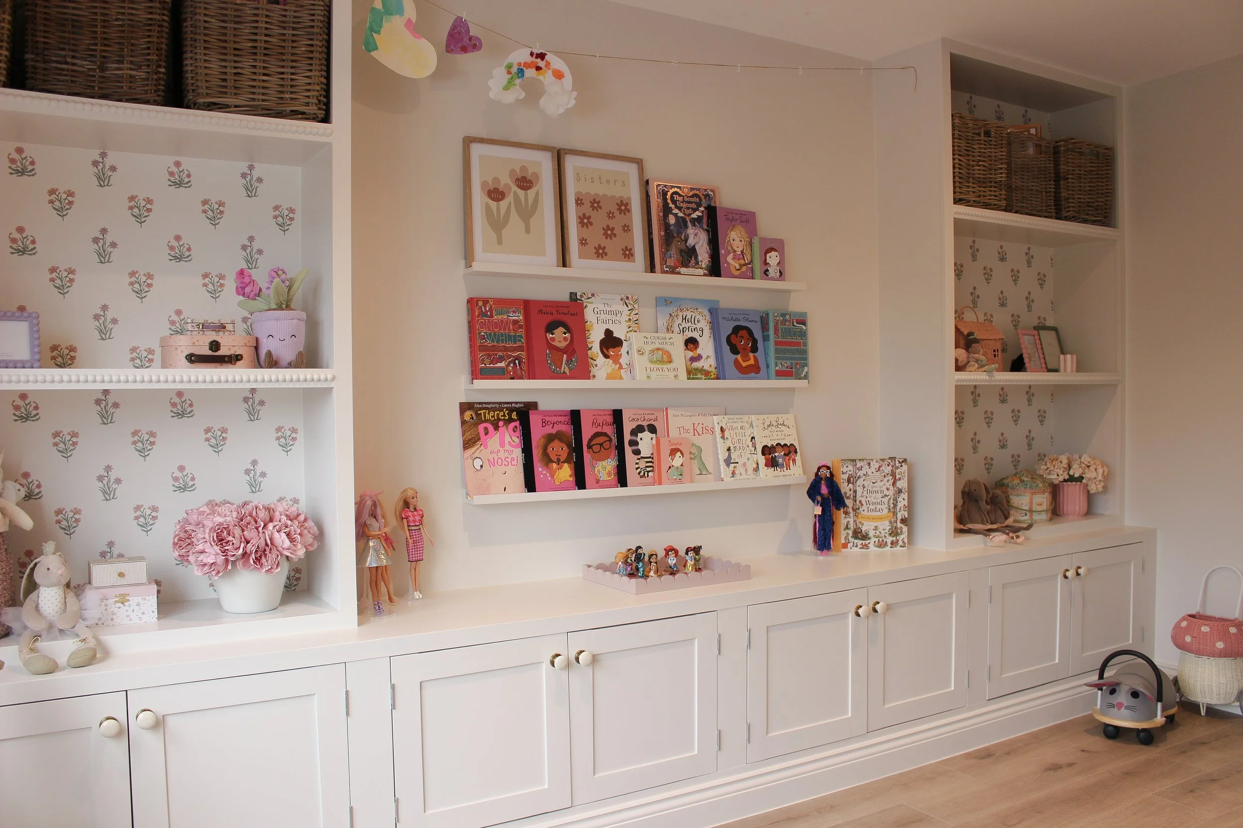 A white bookshelf and cabinet with children's books, toys, and decorative items in a cozy, well lit room. Playroom designed by Yazzmin Lovelle Interiors 