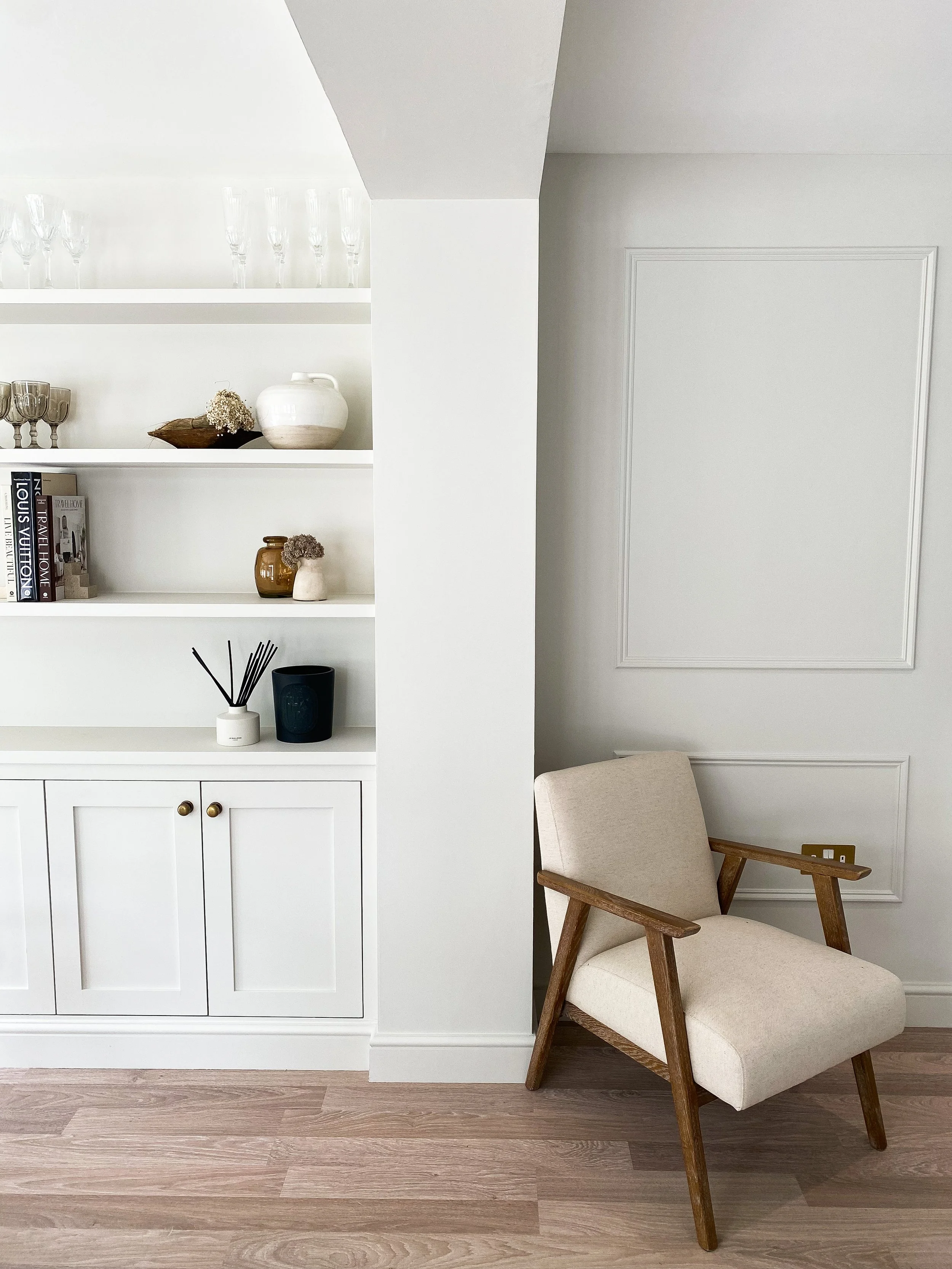 A beige armchair with wooden armrests and legs, positioned next to a white wall with decorative molding, in a bright room with light-colored wooden flooring and built-in white shelving filled with decorative items.
