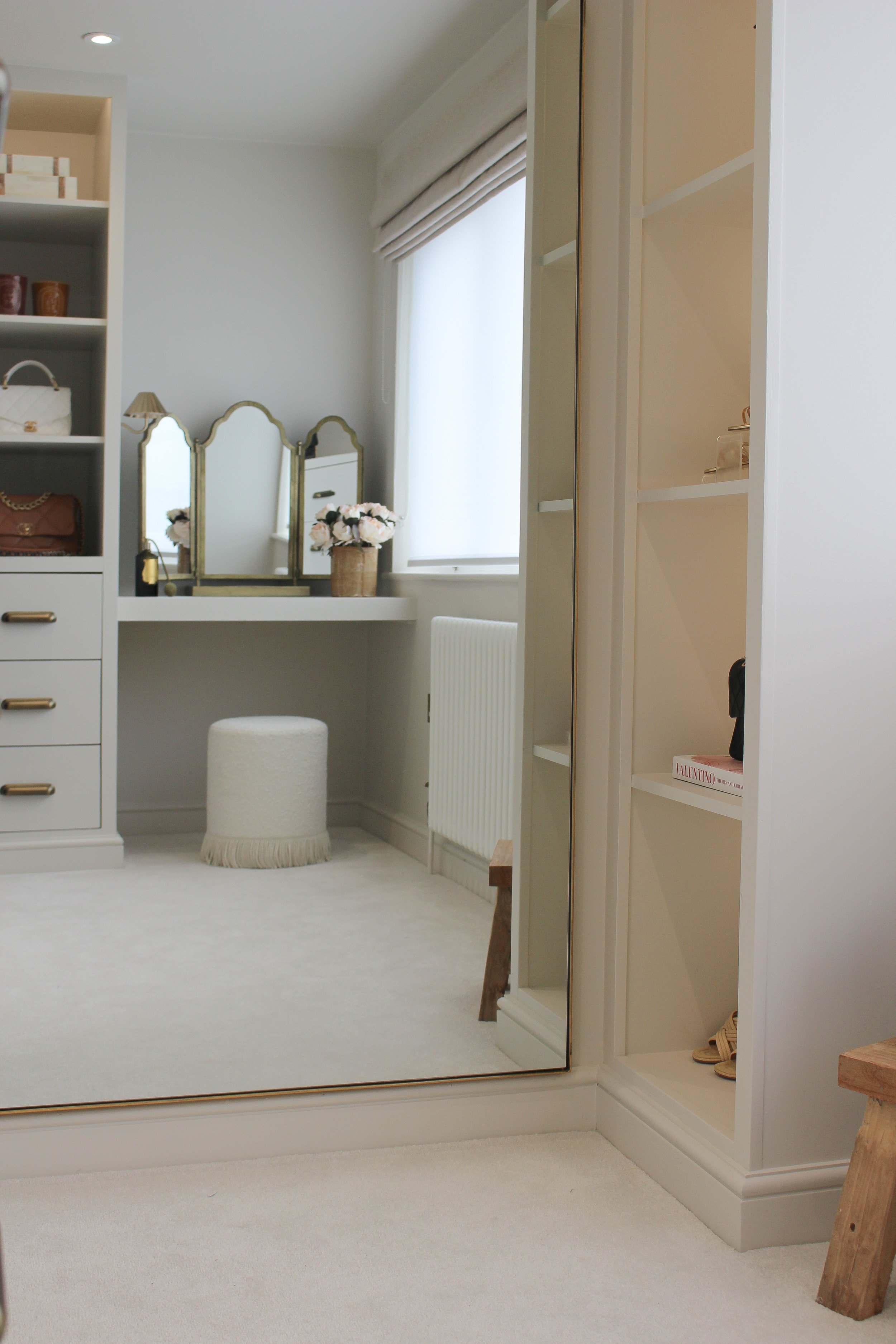 A bright and elegant dressing room with a large mirror, vanity table, potted flowers, shelves with handbags, shoes, and accessories, a small white ottoman, and a window with roman blinds. A room designed by Yazzmin Lovelle Interiors.