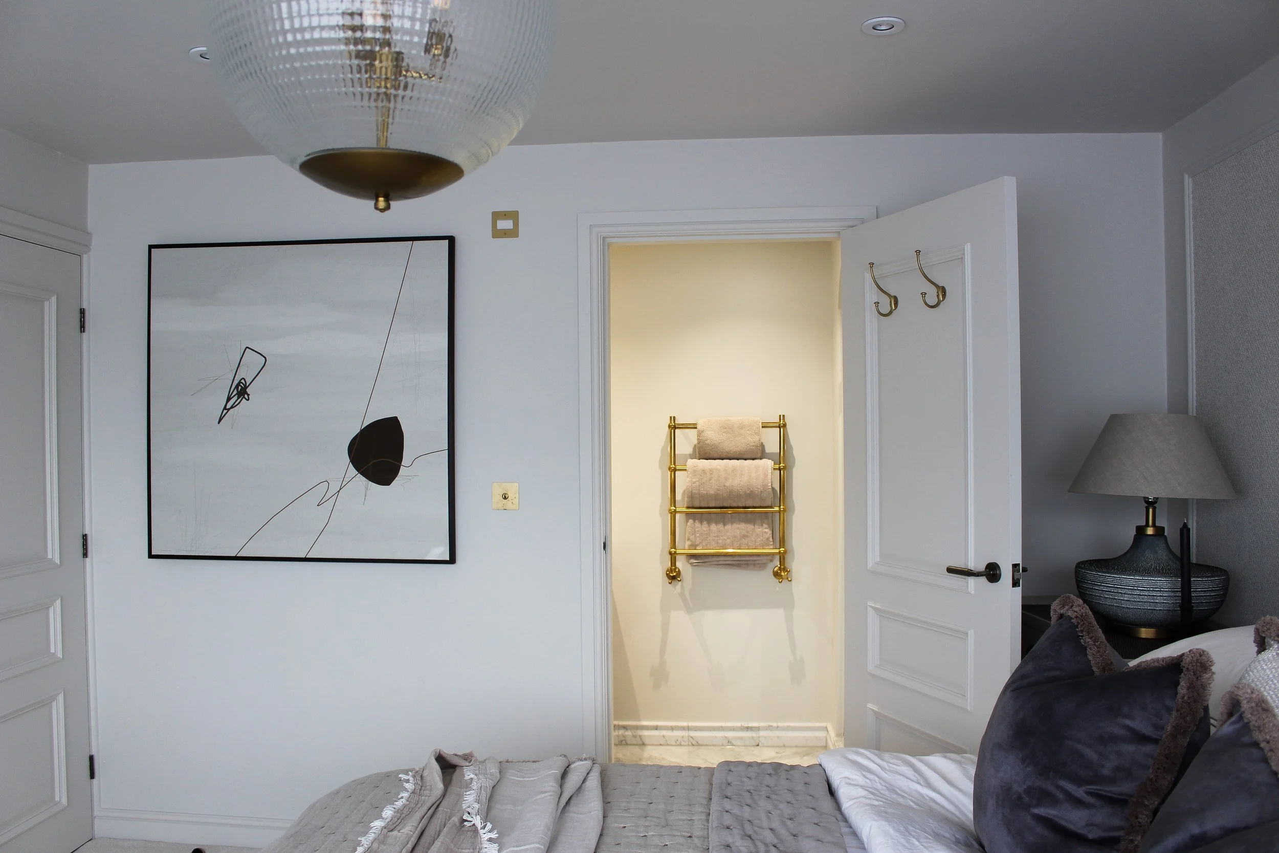 A bedroom with a bed, a lamp on a bedside table and a decorative towel radiator seen through the door leading to an ensuite bathroom with beige walls and a marble shower 