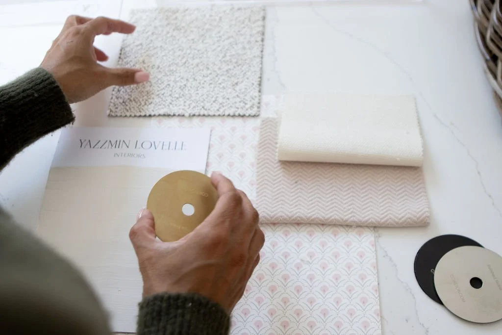 A person arranging fabric and wallpaper samples on a white surface, with several patterned fabric and paint colour swatches, working on a yazzmin lovelle interiors design project