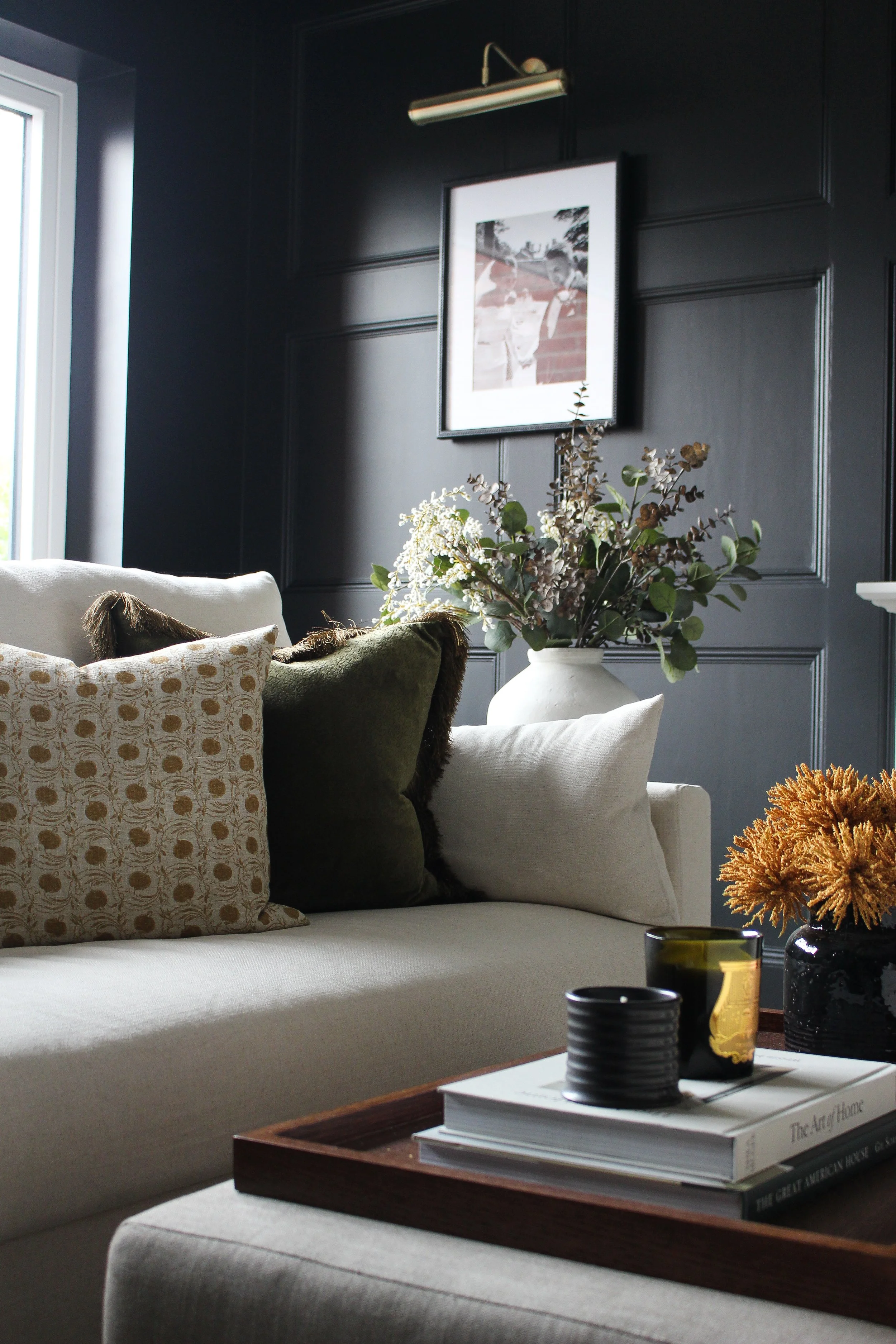 A cozy living room corner with a beige sofa decorated with patterned, solid green, and beige throw pillows. A dark wall with decorative paneling features a framed black-and-white picture, and a modern wall light is above it. A large white vase with g