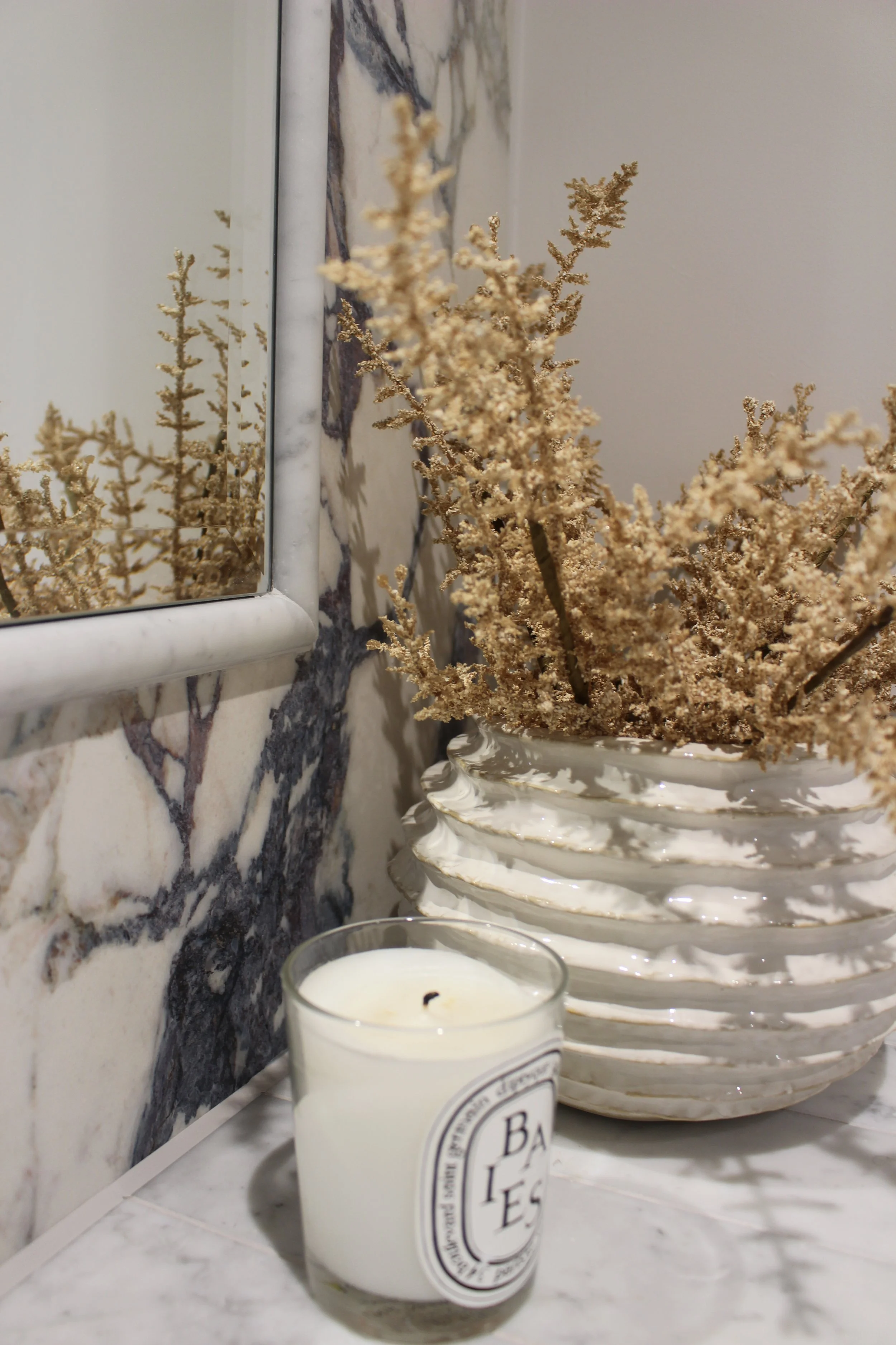 A marble countertop with a white scented candle and a large white ceramic vase filled with dried beige flowers. A mirror on the wall reflects part of the flowers and marble pattern.