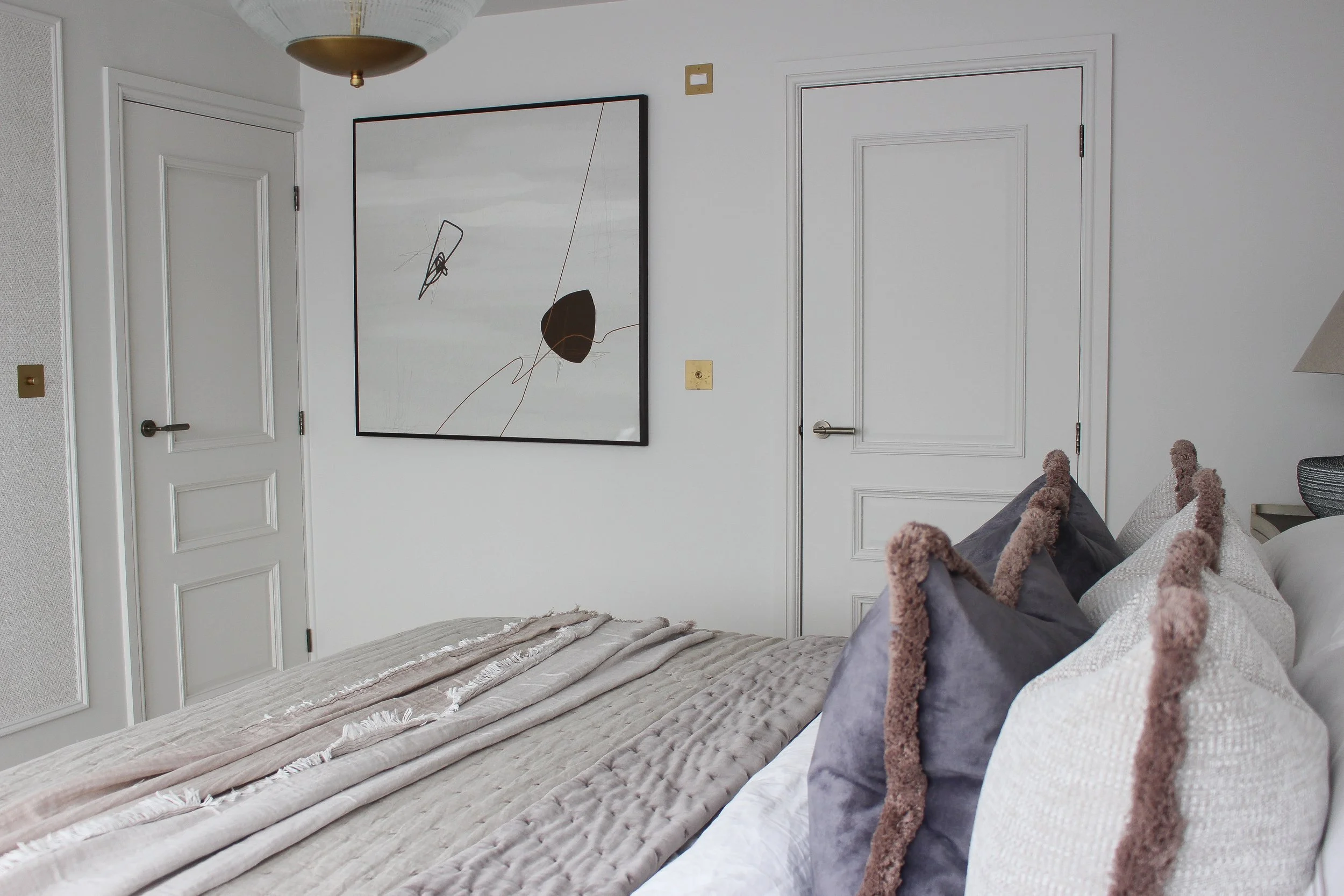 A neatly made bed with beige and white bedding, multiple pillows with fringe edges, in front of white panelled doors and a large modern abstract painting on a white wall in a bedroom. 