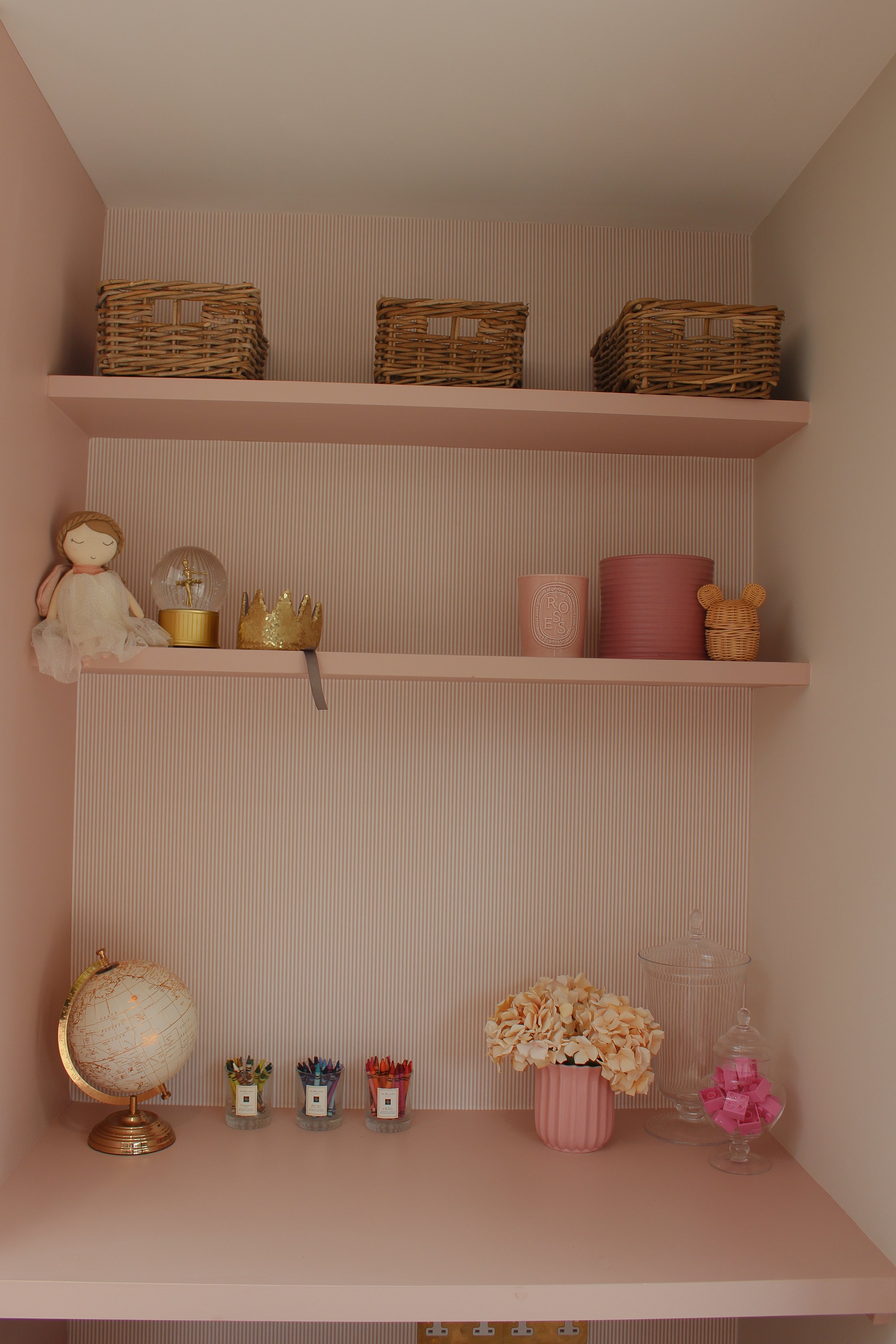 Decorative shelves with woven baskets, a doll, a globe, coloured pencils, flowers, and various decorative items in pink and beige tones. Playroom designed by Yazzmin Lovelle Interiors.