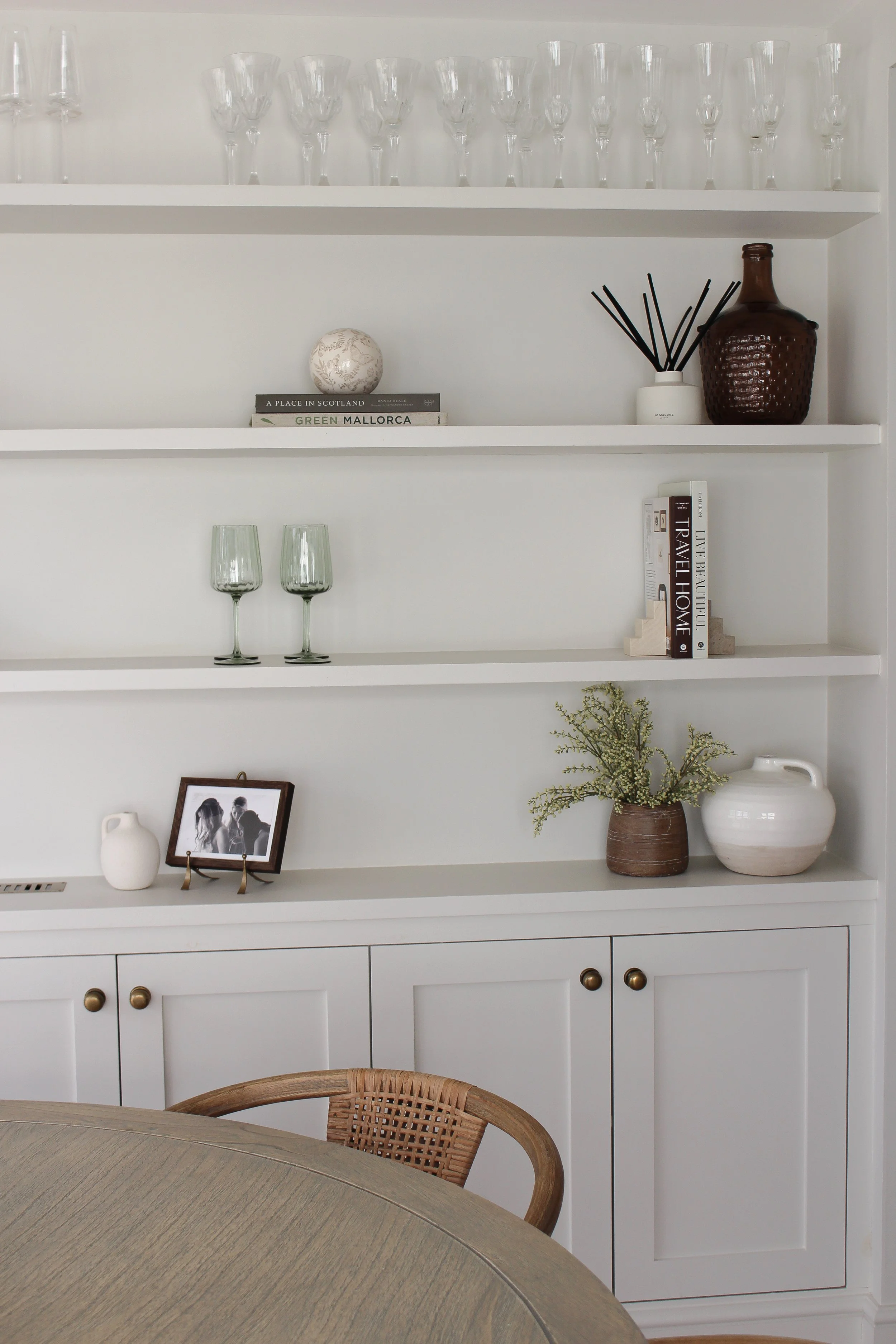 White built-in bookshelf with glassware, books, a reed diffuser, decorative vases, a framed black-and-white photo, and a plant, with a wooden chair and table in front. Dining area designed by Yazzmin Lovelle Interiors.