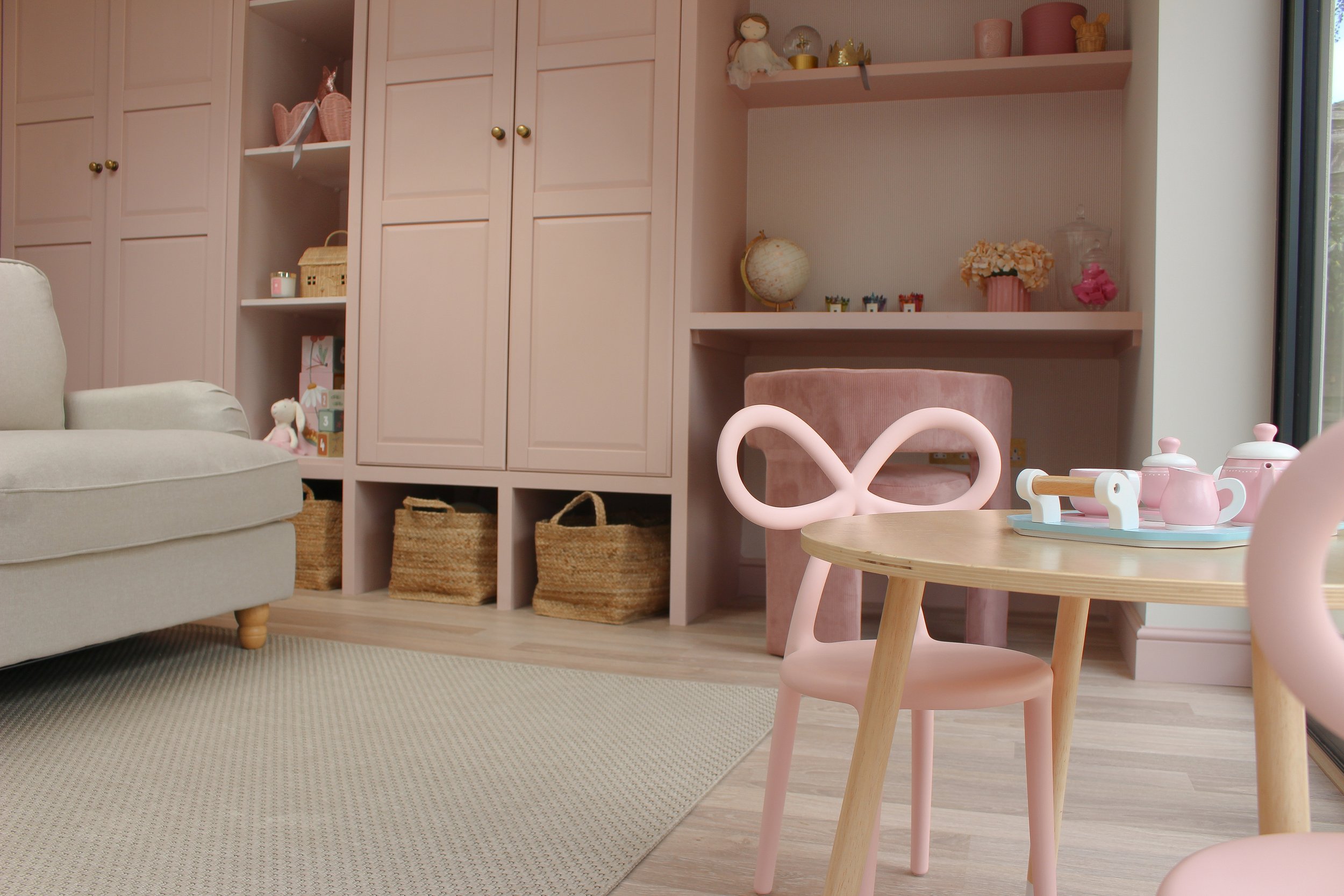 A cozy child's playroom with a pink and beige color scheme. Contains a pink chair with bow back detail, a small round table with a pink tea set, a beige sofa, built-in storgae with toys and decor, and a large window. Playroom design 