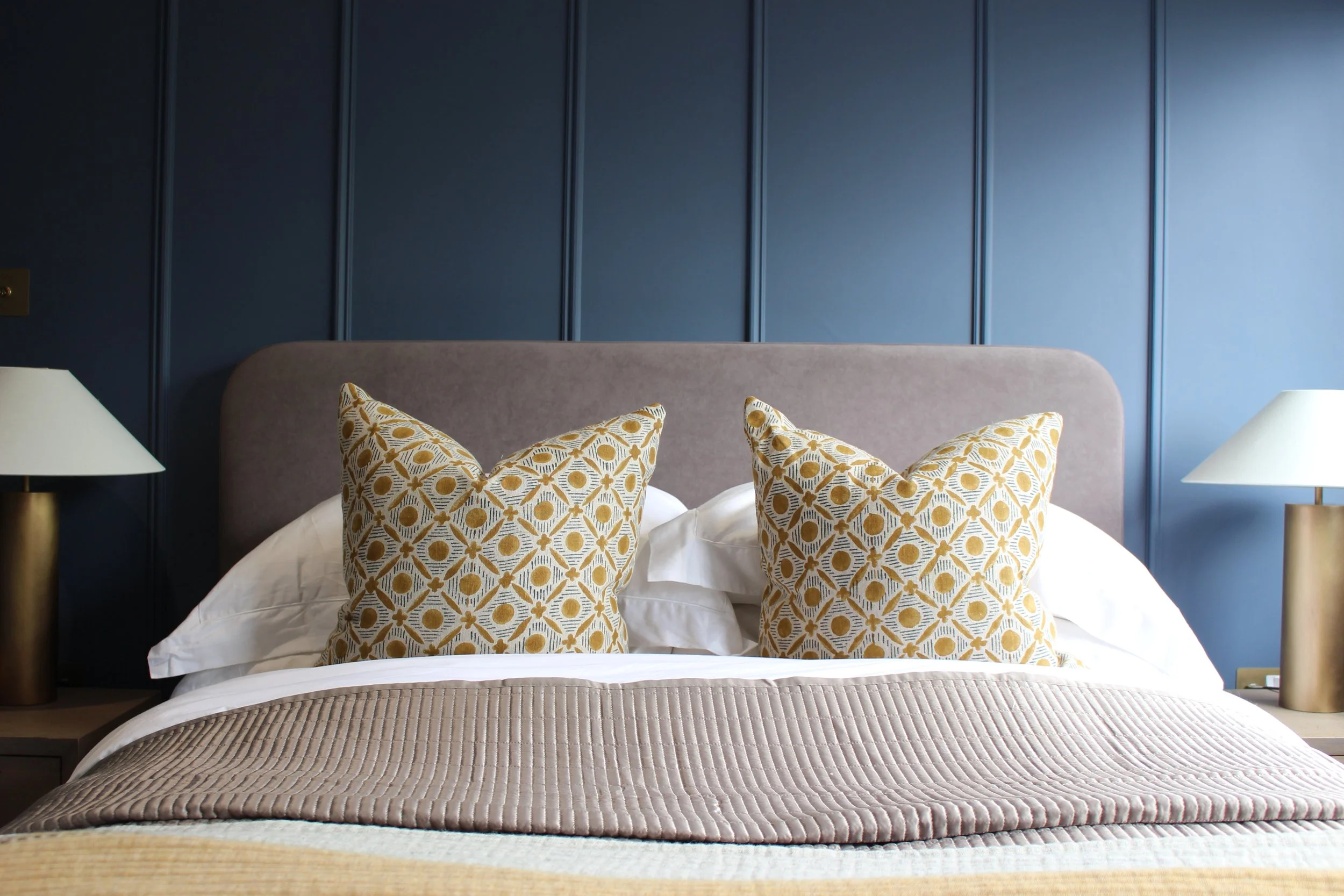 A neatly made bed with two decorative pillows featuring a yellow and white pattern, grey bedspread, in a bedroom with blue panelled walls and two white lamps with gold bases. Guest Bedroom designed by Yazzmin Lovelle Interiors.