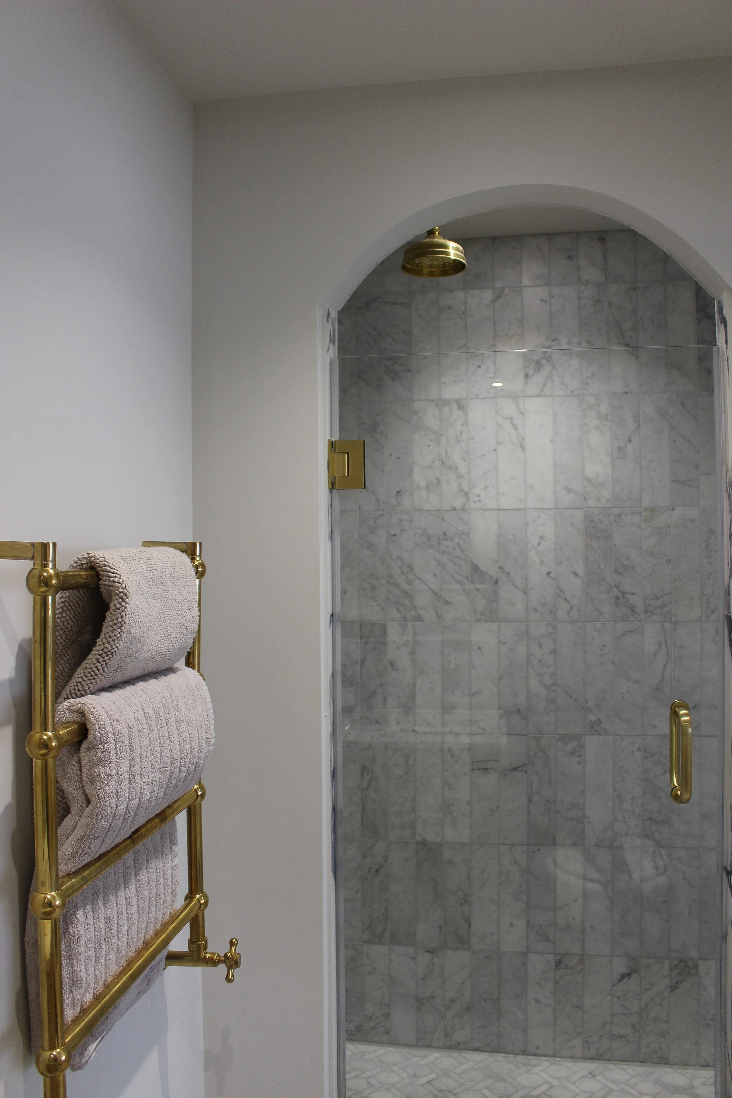 A bathroom with a marble shower, antique brass fixtures, and a towel radiator holding beige towels. Master bedroom designed by Yazzmin Lovelle Interiors