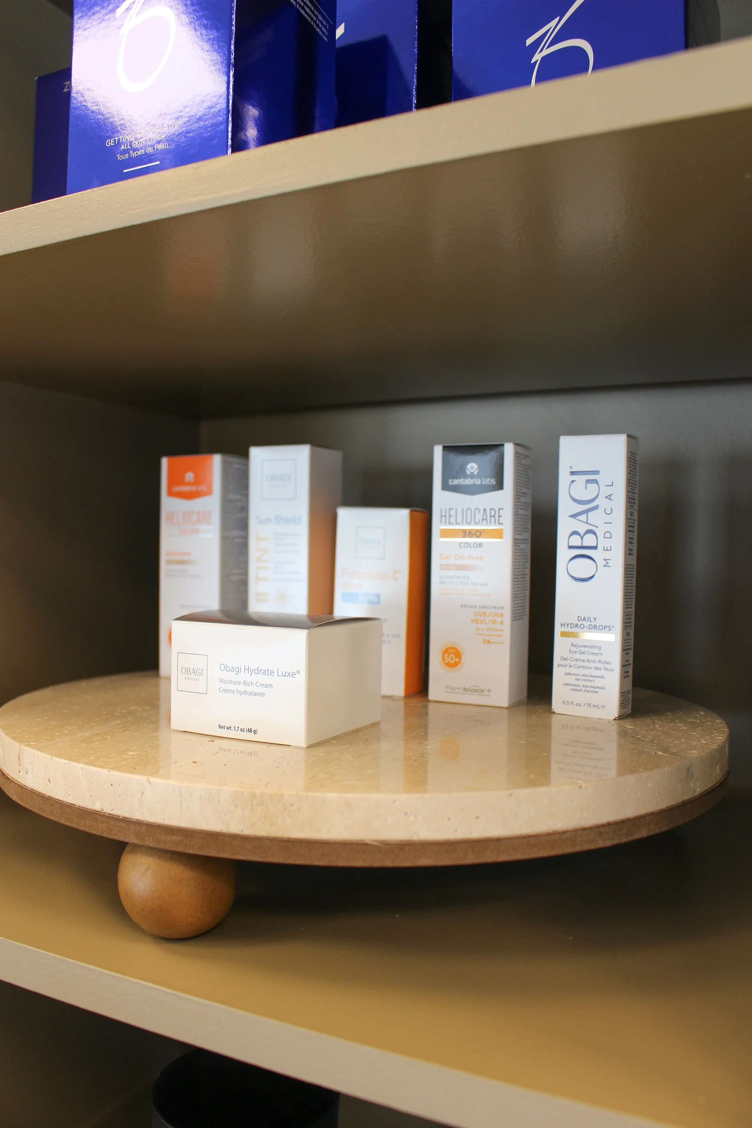Various skincare products arranged on a small round beige table, shelves visible above and below, including Obagi Hydrate Luxe, Helocare 360, Obagi Daily Hydro-Drops, and other sun protection and skincare creams.