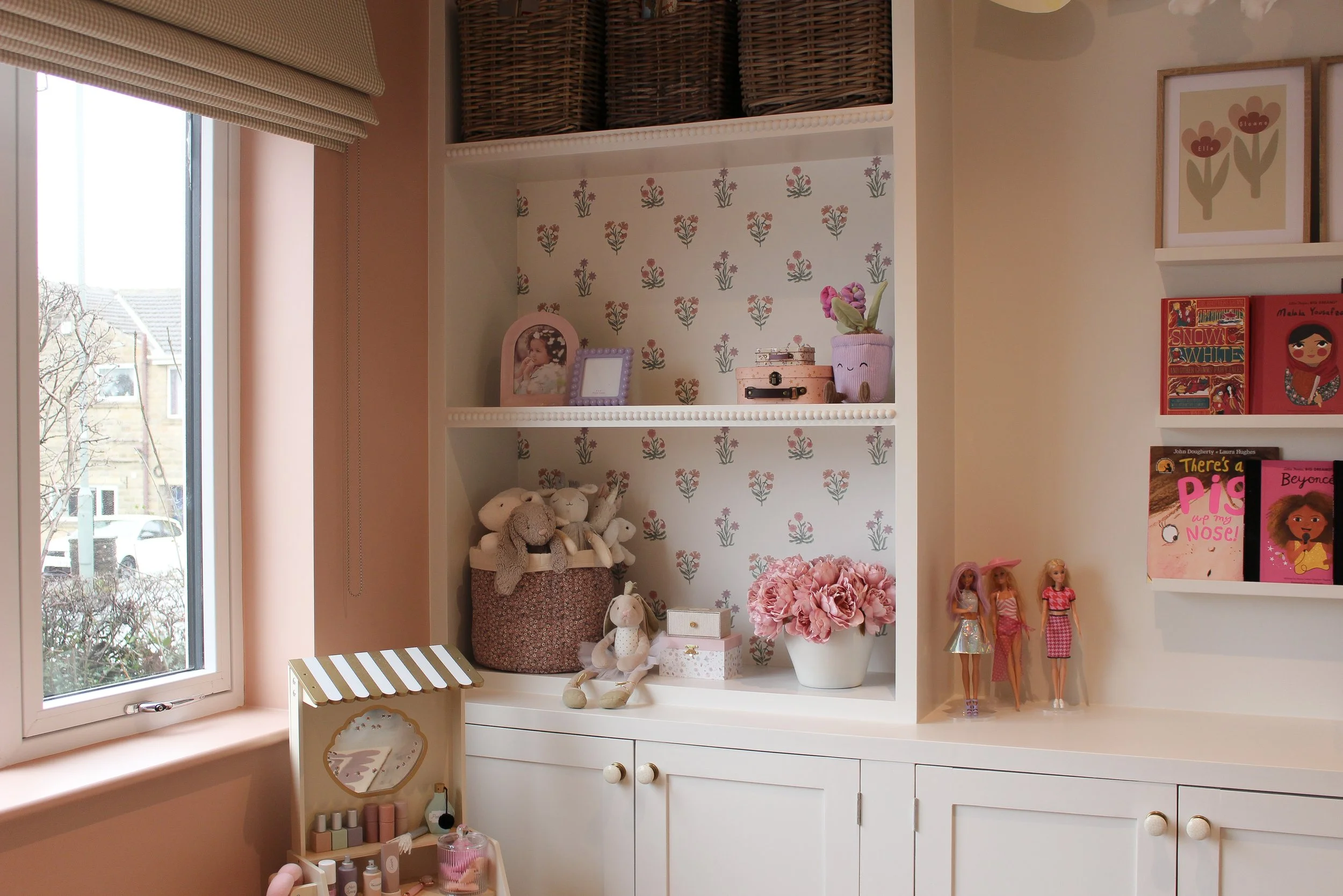 Decorated playroom shelf with plush toys, framed photos, pink flowers in a vase, children's books, and dolls, next to a window with a gingham blind. Guest Bedroom designed by Yazzmin Lovelle Interiors.