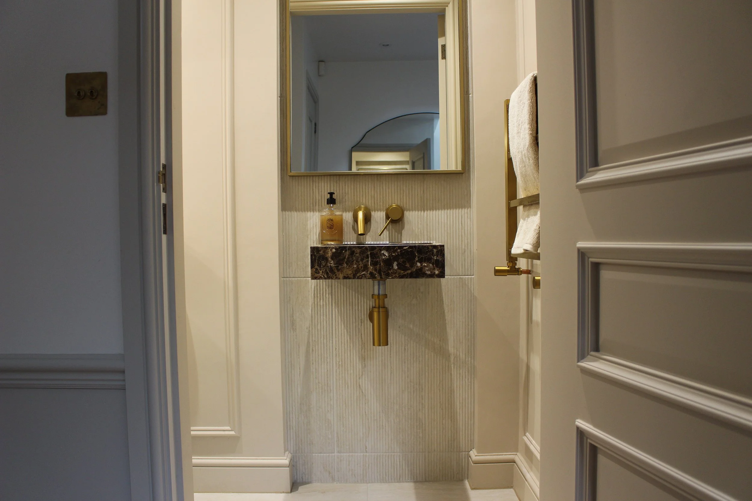 A small downstairs WC with a statement marble sink, gold fixtures, a mirror, towel radiator, and beige coloured panelled walls with tiling. Downstairs toilet designed by Yazzmin Lovelle Interiors.