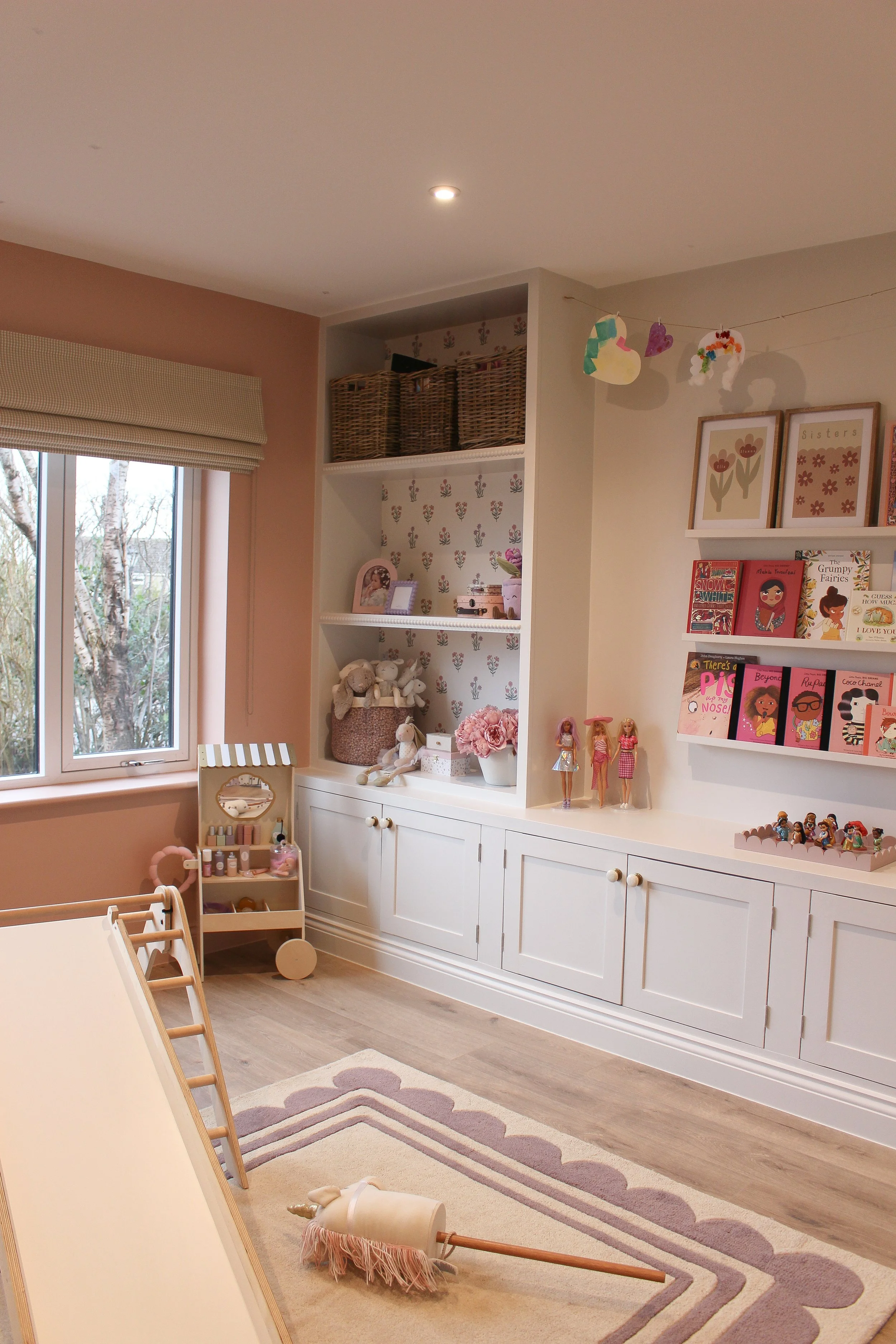 A bright, children's playroom with a window, white built in shelves filled with toys and books, and a lilac and beige rug on wooden flooring. Toys, dolls, and decorative items are arranged throughout the room. 
