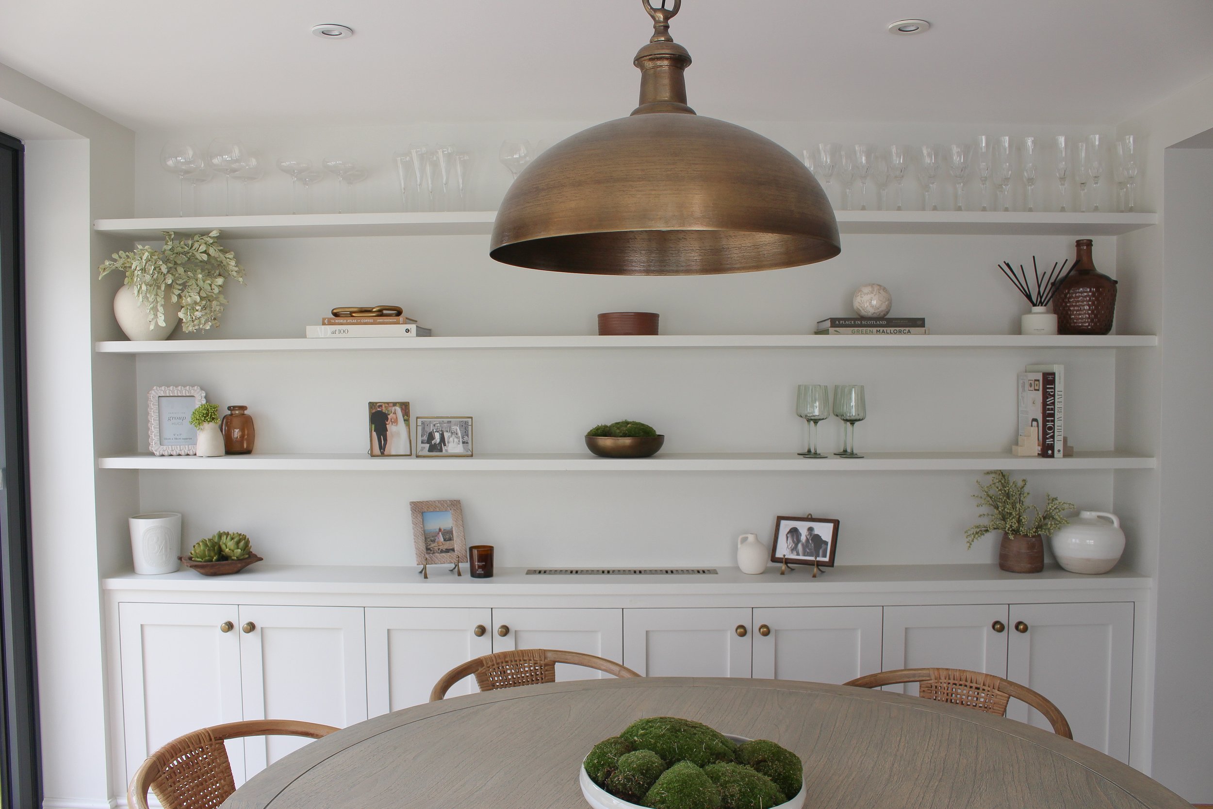 A dining room with a white built-in shelf displaying decorative items, photos, and books, a large bronze pendant light overhead, and a wooden dining table with a bowl of green moss at the center. Dining area designed by Yazzmin Lovelle Interiors.