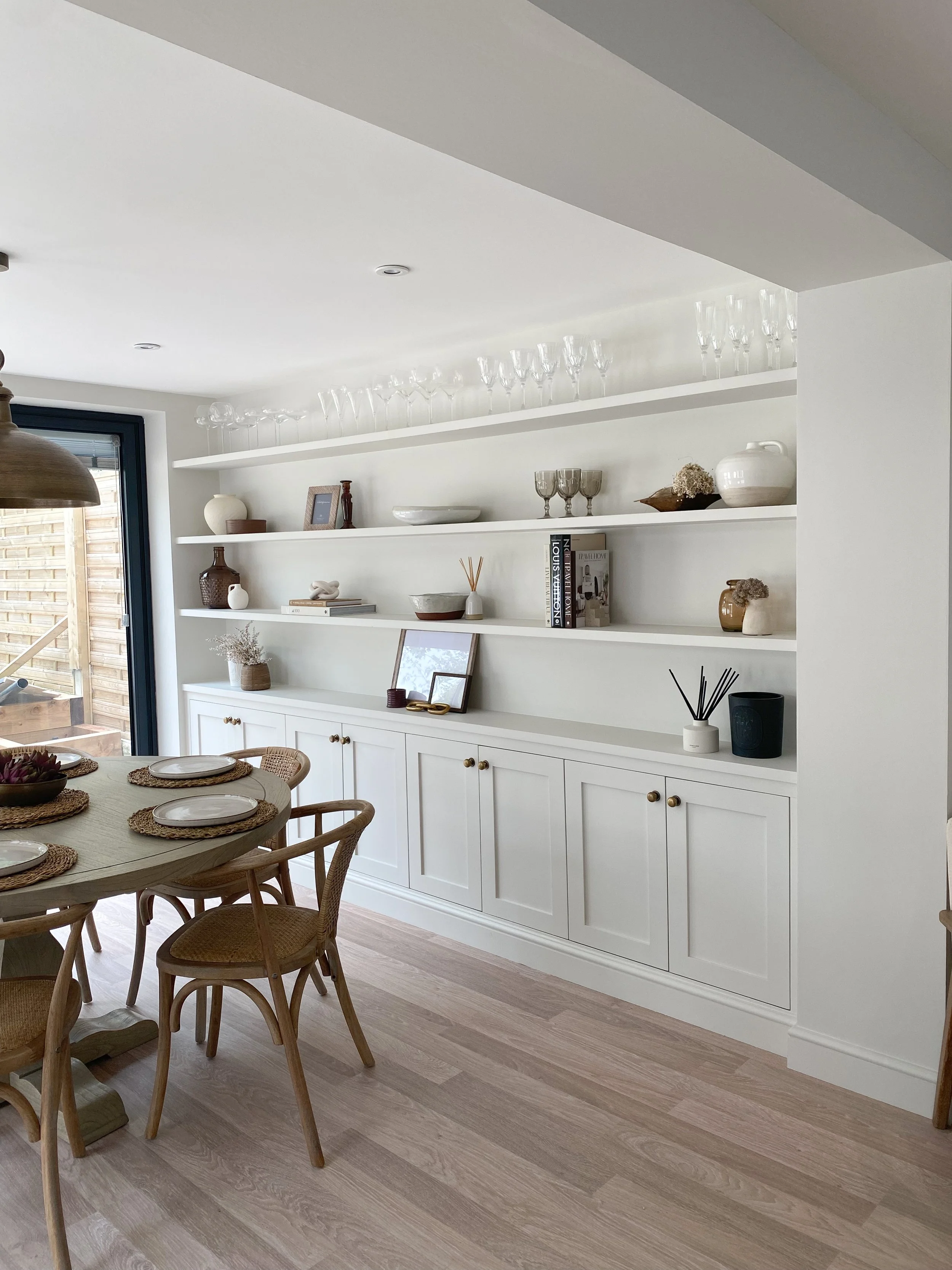 A dining room with a white built-in cabinet, open shelves with decorative vases, books, and glassware, a wooden dining table set with plates and placemats, and natural light coming through a glass door.
