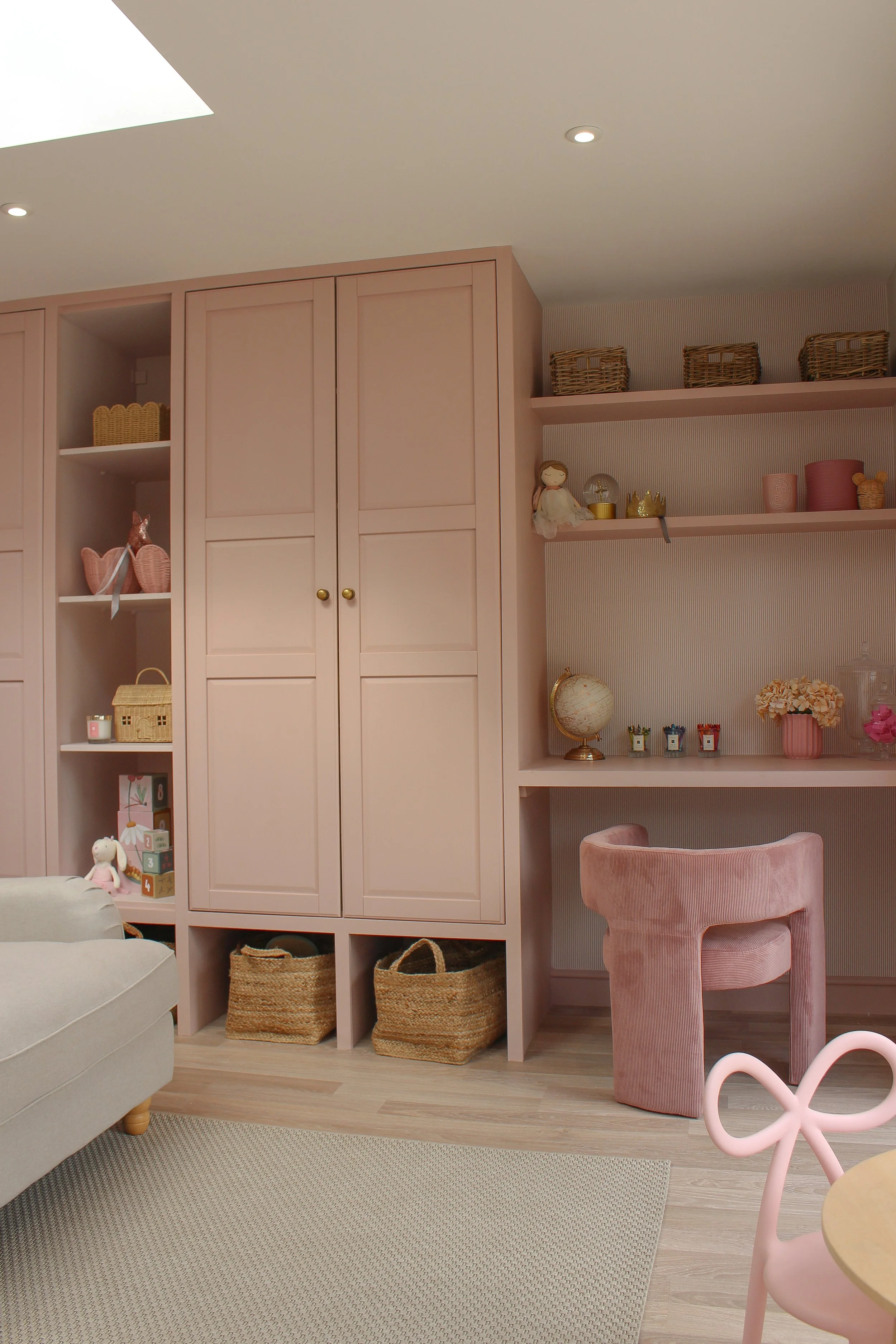 Pink playroom, featuring a built in cabinets, open shelves with decorative items, a pink velvet chair, and a desk with small plants and a globe. Playroom designed by Yazzmin Lovelle Interiors.