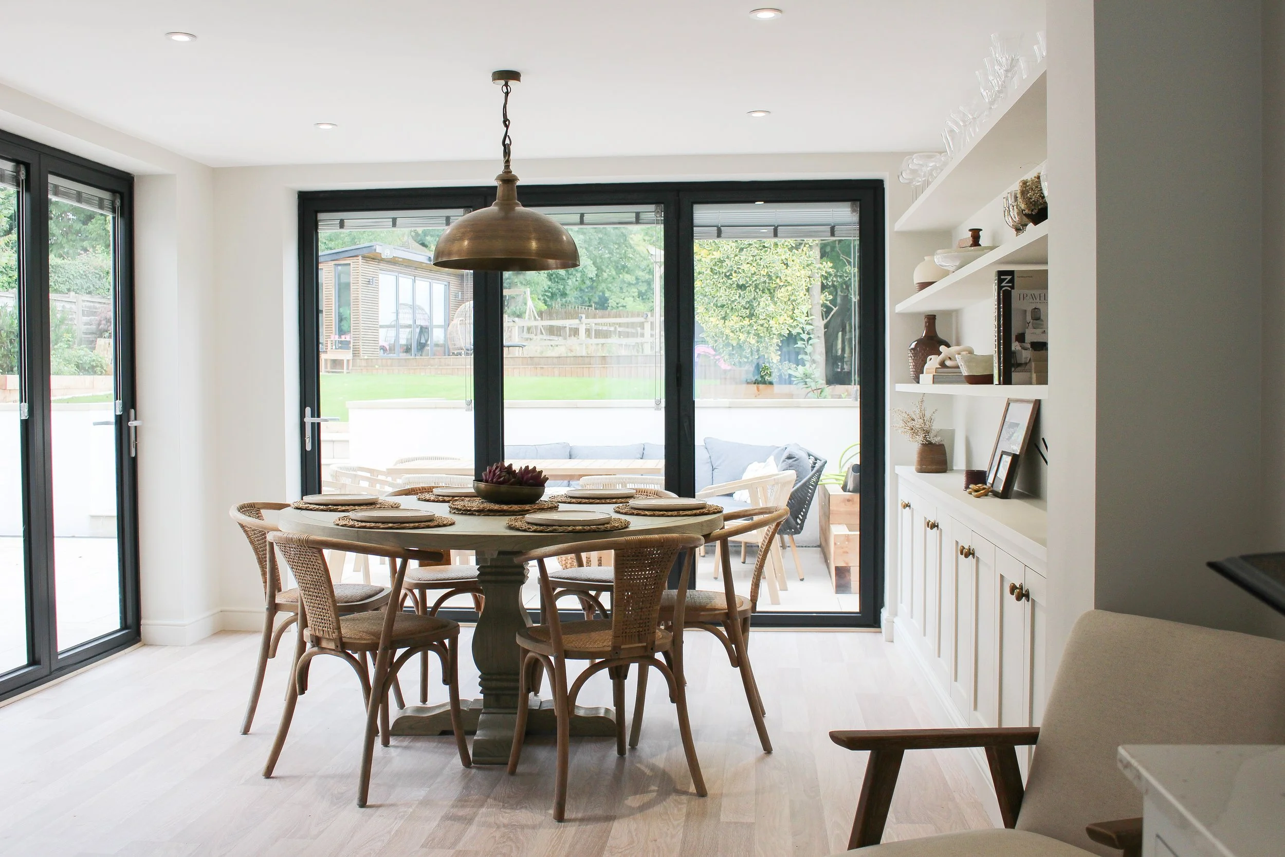 Bright dining room with a round table set for meals, surrounded by wicker chairs, large glass sliding doors leading to an outdoor patio with seating, white built-in shelves with decorative items, and light wood flooring.