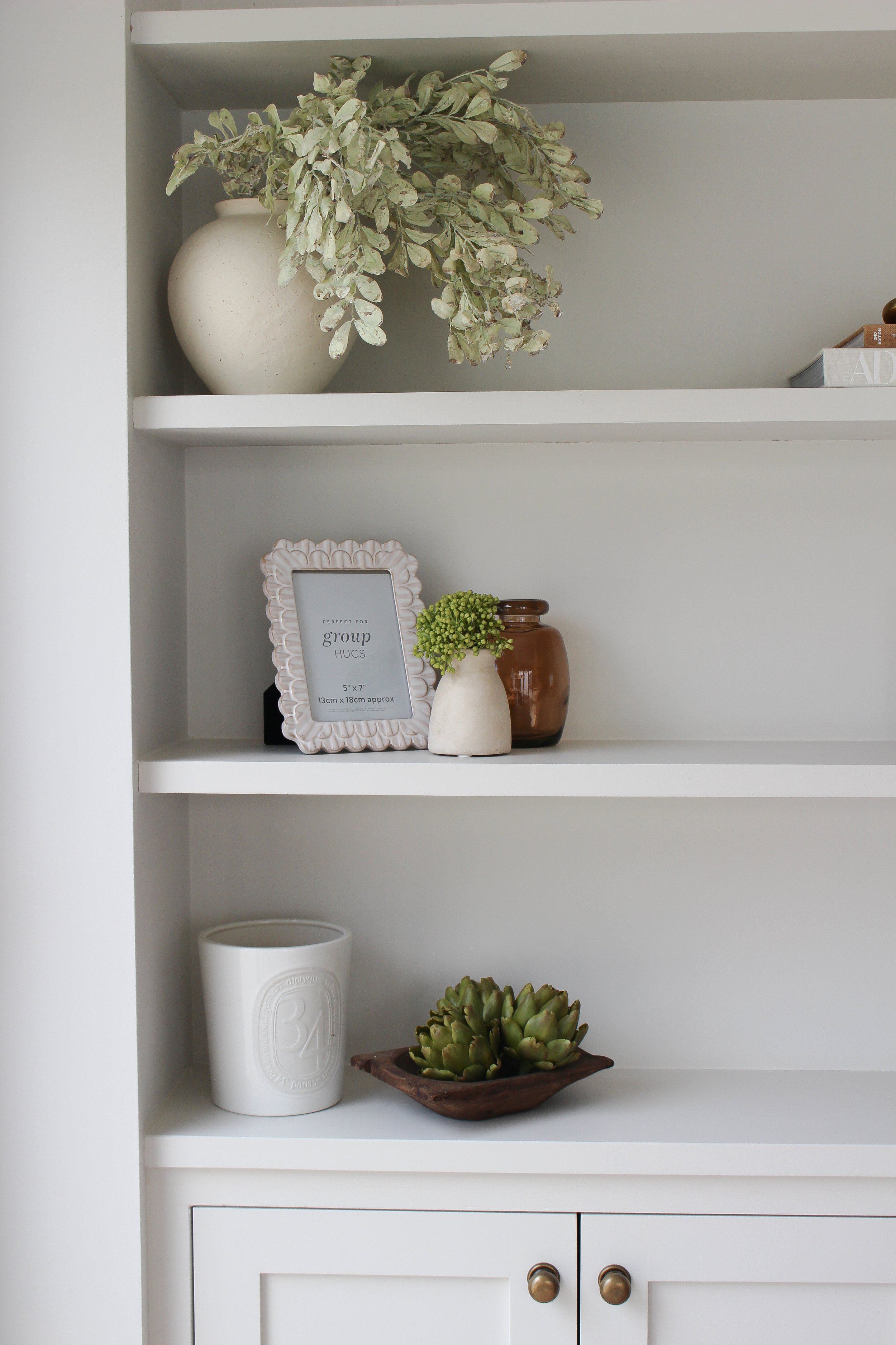 A white bookshelf with decorative items including a large white vase with greenery on top, a small white vase with green flowers, a brown jar, a white framed photo, a white ceramic pot, and a small wooden dish with succulents.