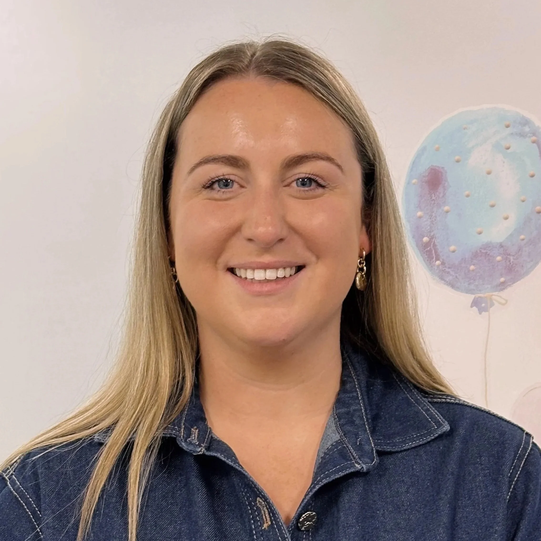 A smiling woman with blonde hair, wearing a denim jacket, standing in front of a white wall with a pastel-colored balloon.