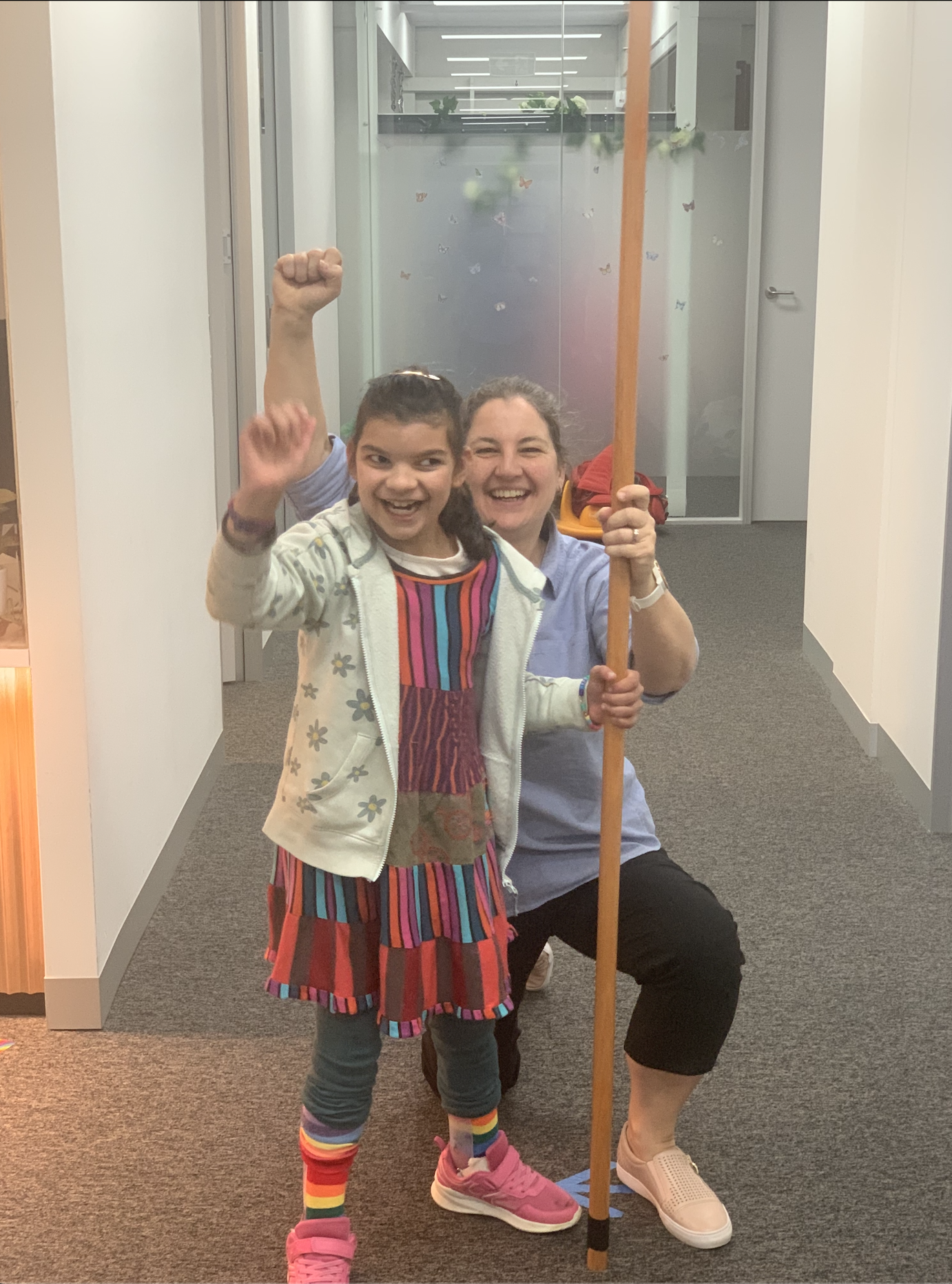 A young girl and an adult woman in an indoor hallway, celebrating and smiling. The girl raises one fist in victory, dressed in a colorful striped outfit and rainbow socks. The woman holds a long orange staff and is kneeling.