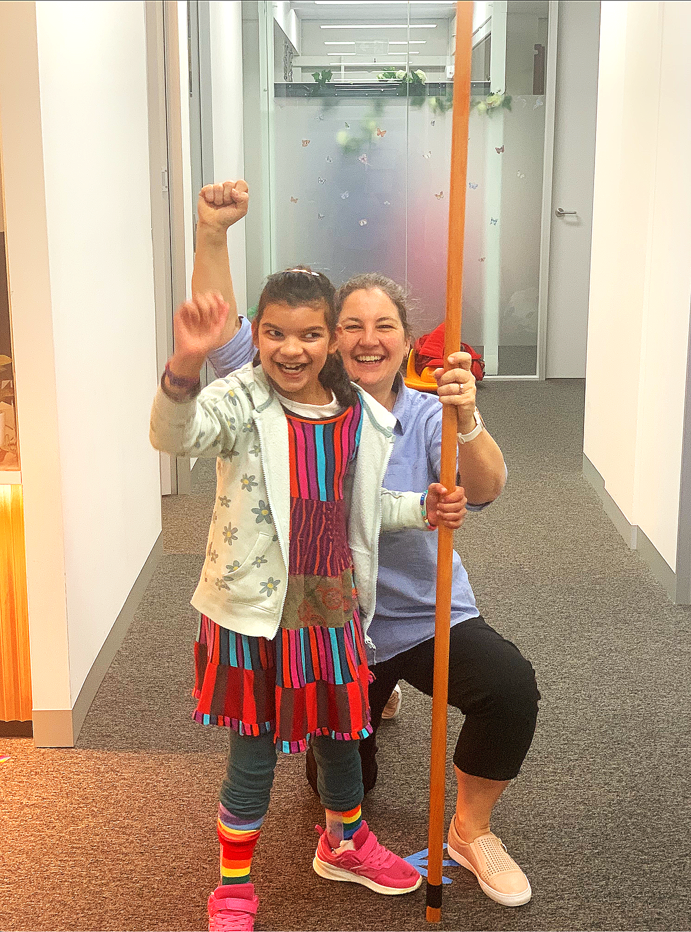A young girl smiling happily with a woman, both raising their fists and holding a staff in a hallway with glass doors and butterfly decorations.