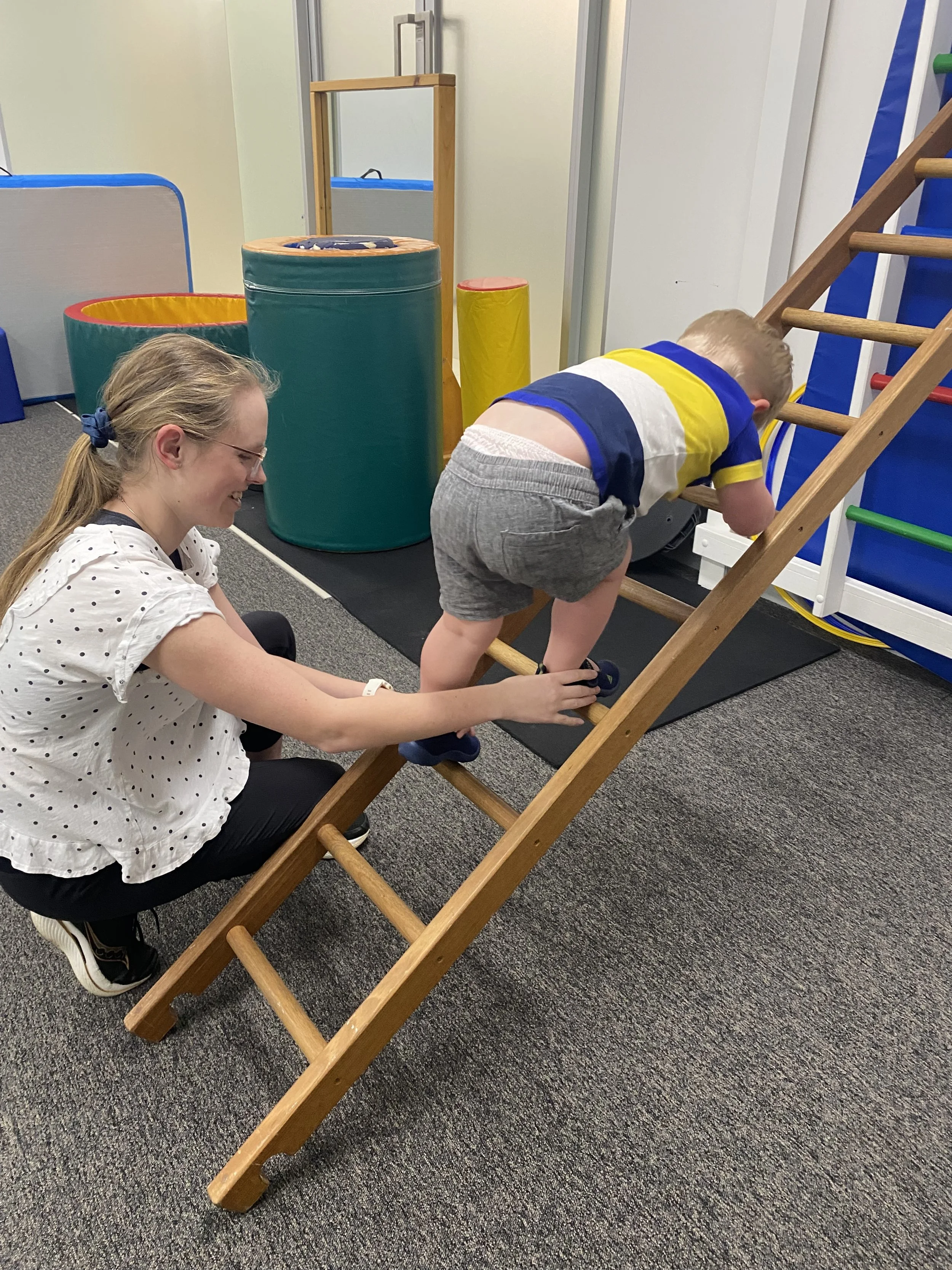 A young boy climbing up a small wooden ladder in a play area, with a woman crouching beside him helping. The room has colorful padded play equipment, including a blue and yellow slide, and soft cushioned mats.