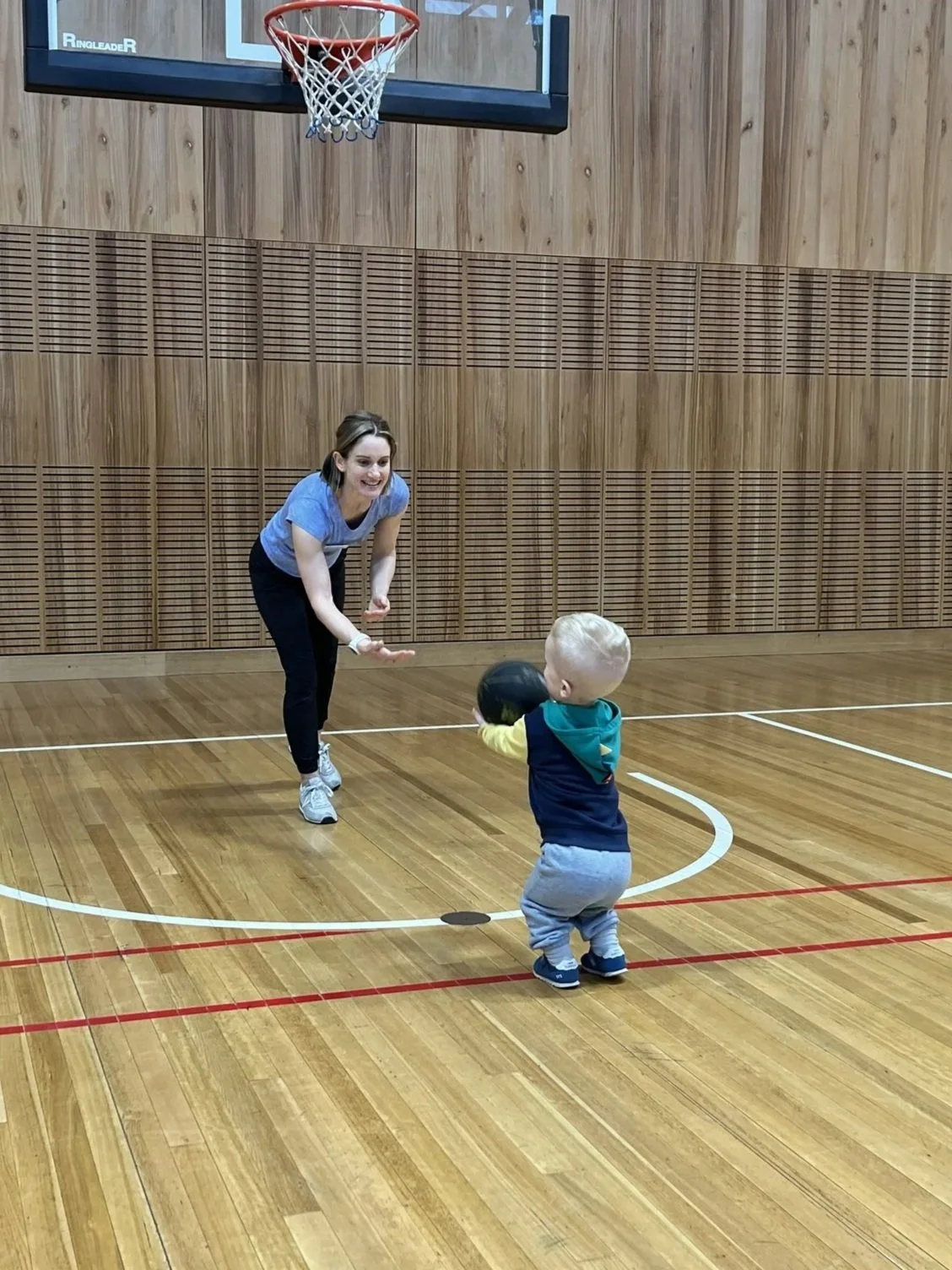A woman playing basketball with a young child in an indoor gymnasium with wooden floors and walls.