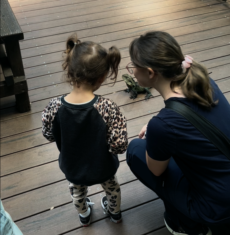 A young girl with two ponytails and a woman with glasses squatting on a wooden deck, observing a small lizard.