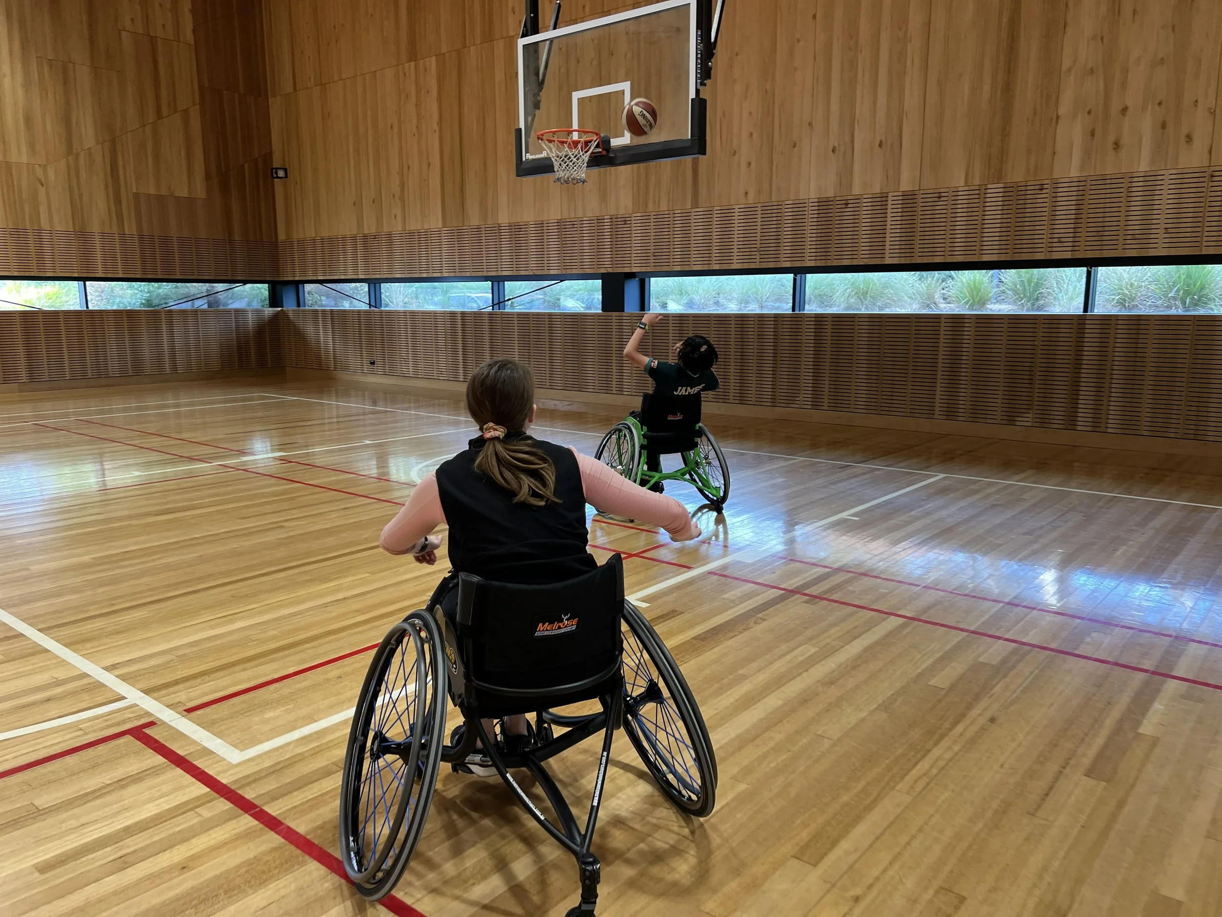 Two women in wheelchairs playing wheelchair basketball in an indoor gymnasium with wooden floors, walls, and large windows showing greenery outside.