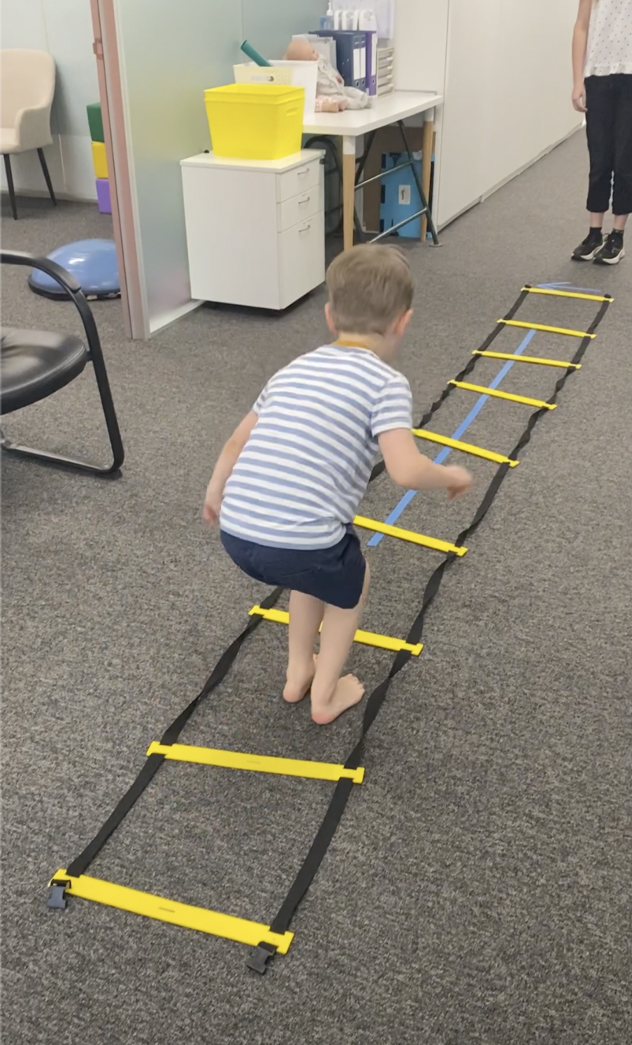 Child crawling on a yellow and black agility ladder on carpeted floor in an indoor room, with a chair, a white cabinet, and a table with supplies in the background.