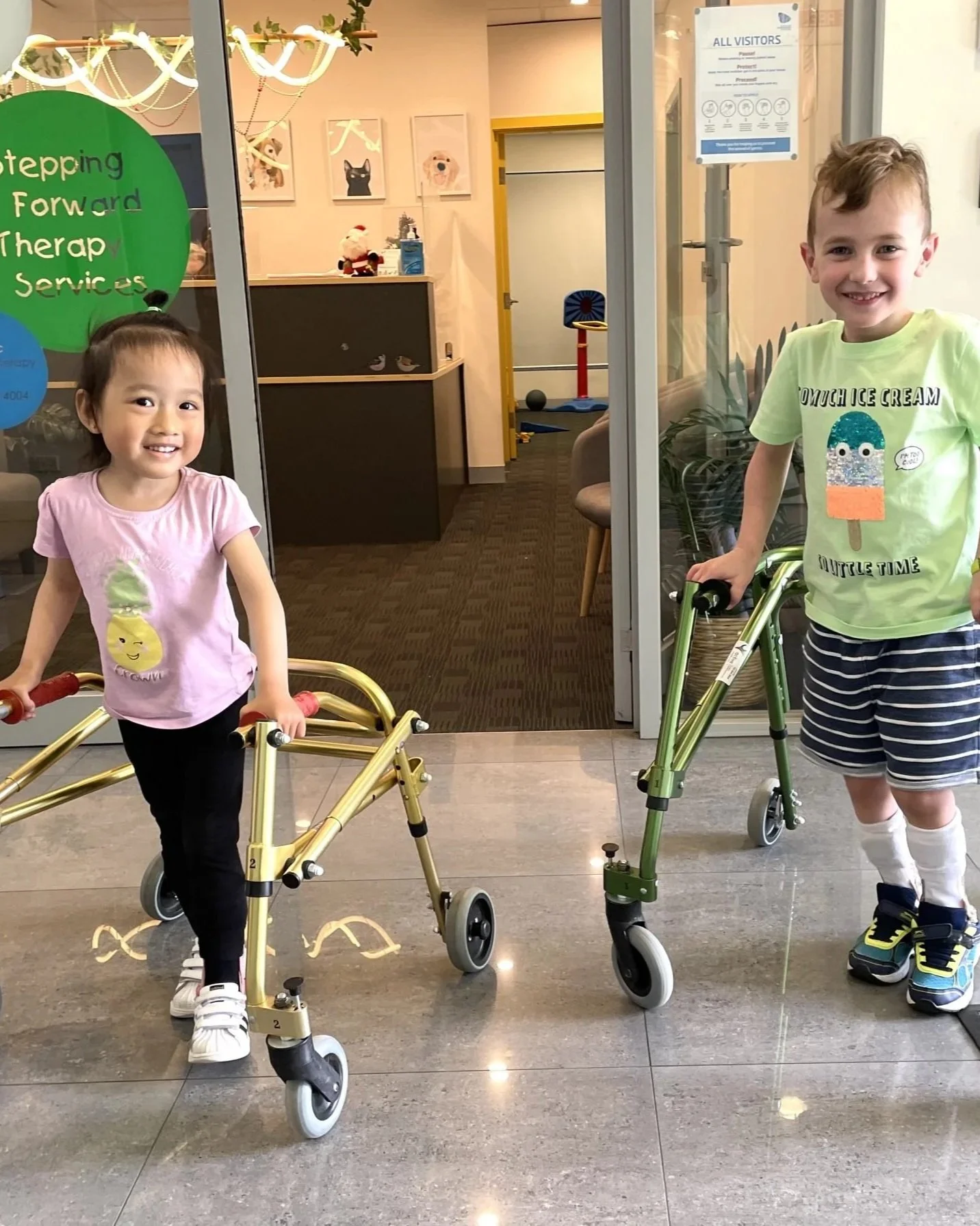 Two young children, a girl and a boy, smiling and holding onto walkers inside a building. The girl is wearing a pink shirt and black pants, and the boy is in a green shirt and striped shorts. A decorated lobby or reception area is visible in the background.