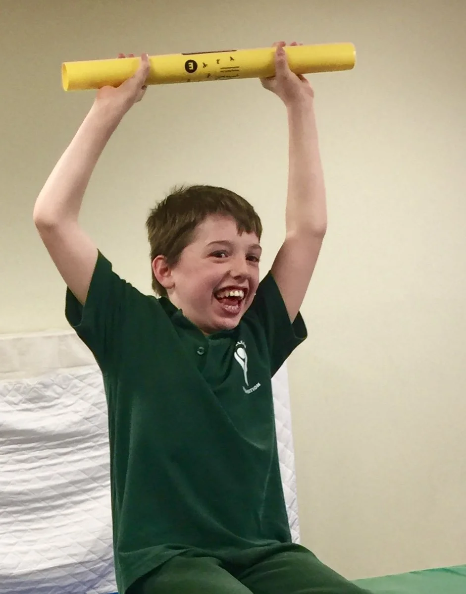 A smiling boy holding a yellow pipe above his head indoors.